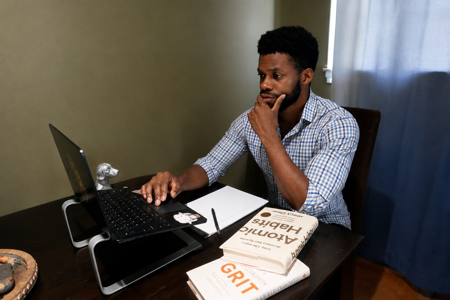 A man sitting at a desk, looking at a laptop, with a notebook, pen, and books on the desk, in a room with a window and curtains.