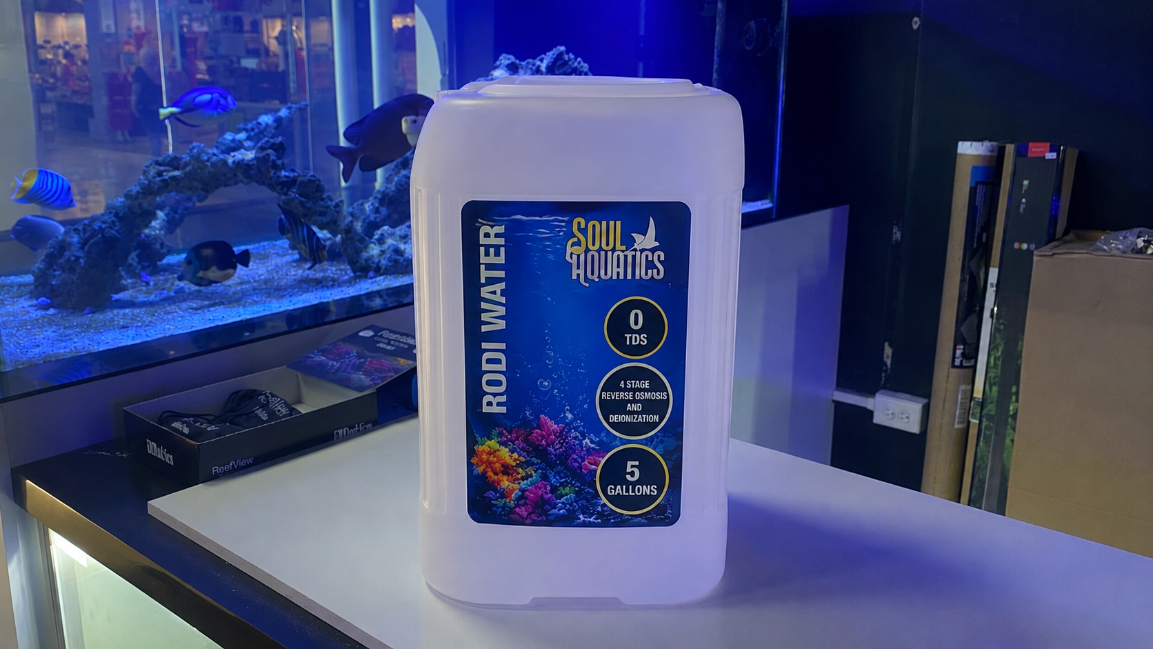 Container of Soul Hydraulics Rodi Water with 0 TDS, 5 gallons, on a white surface, with an aquarium and store shelves in the background.