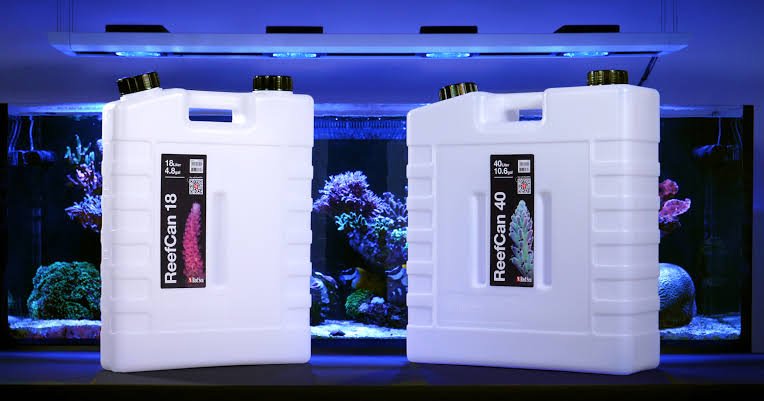 Two large white containers labeled ReefCoan with coral images, placed in front of a vibrant aquarium filled with various colorful corals and marine life.