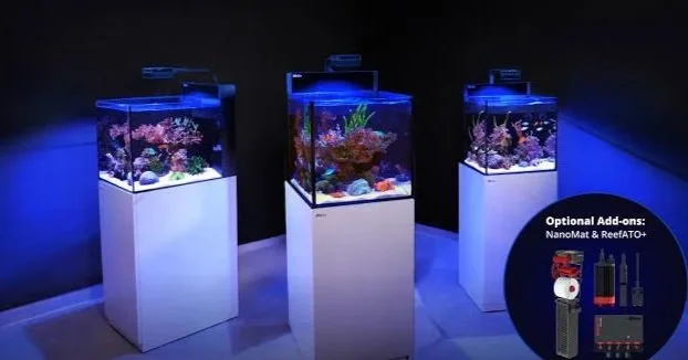 Three illuminated saltwater aquariums with colorful coral and fish on white stands, in a dark room, with an inset showing optional equipment.