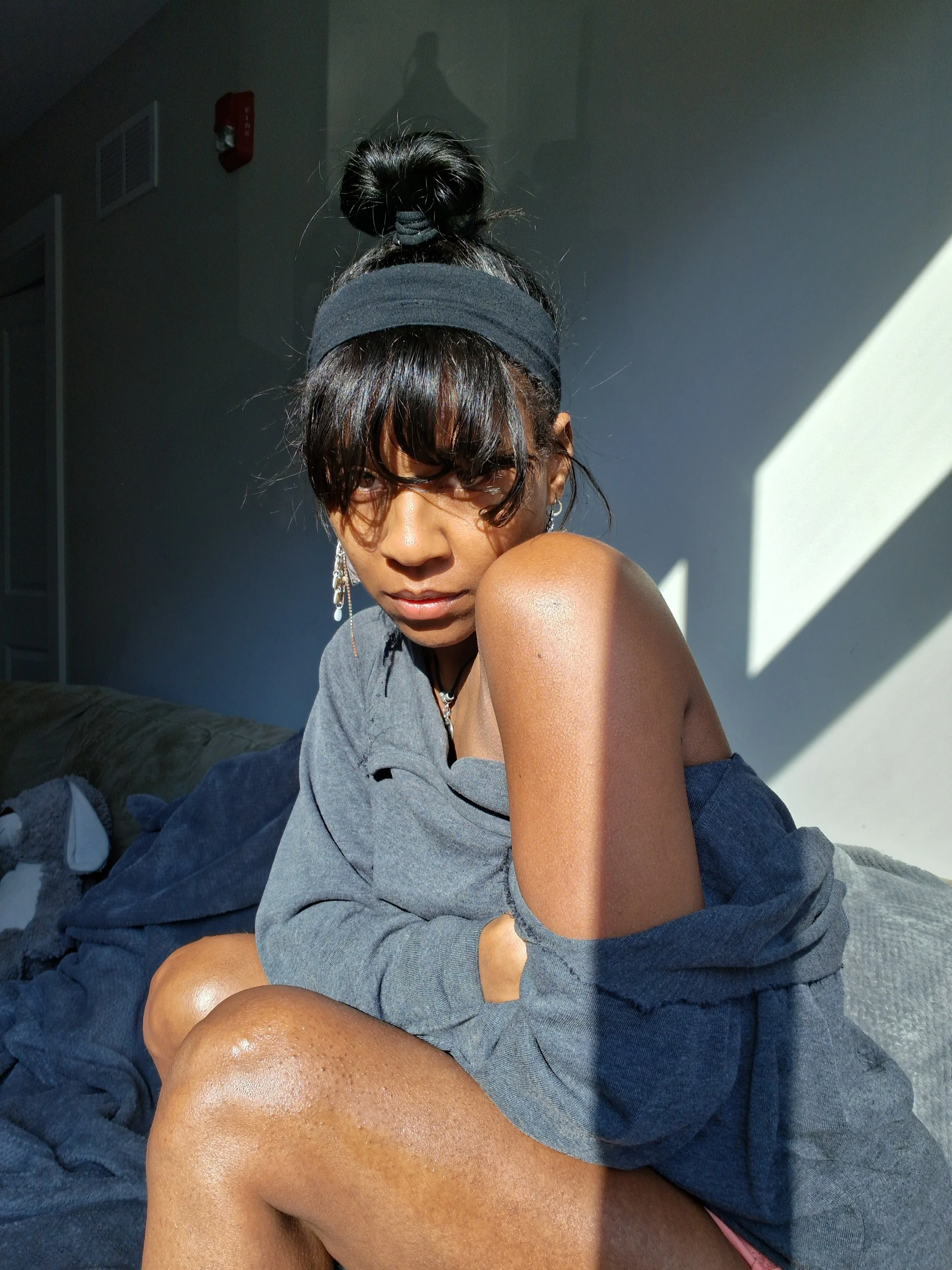 A woman with dark hair tied in a bun, wearing a black headband and earrings, sitting on a bed in a room with sunlight casting shadows on the wall. She is partially covered with a gray hoodie and pink clothing, posing for the photo.
