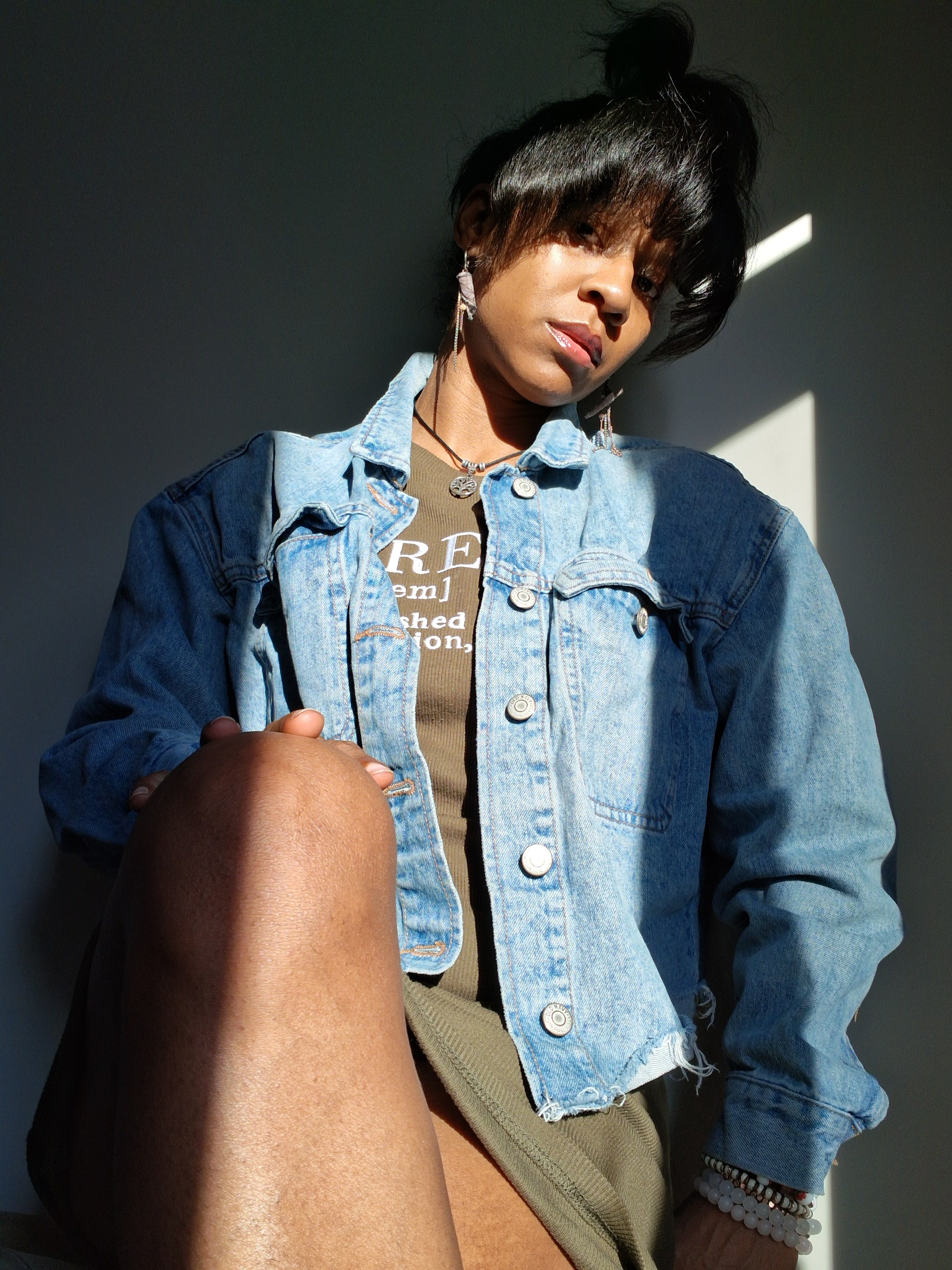 A young person with dark, styled hair and earrings, wearing a denim jacket and a brown t-shirt, poses indoors with dramatic lighting and shadow effects.