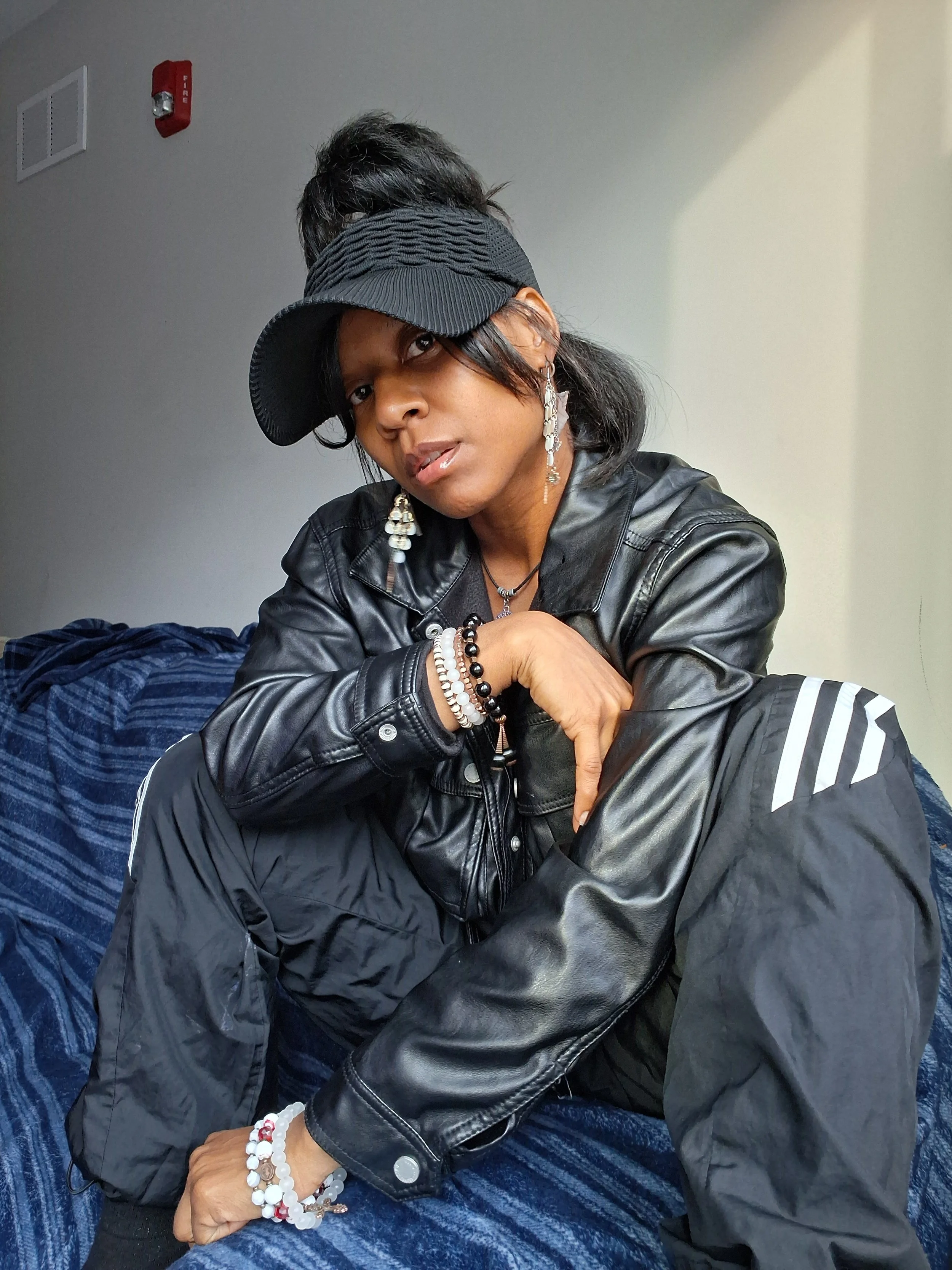 A woman with dark hair, wearing a black leather jacket, multiple beaded bracelets, and earrings, sits with her arms crossed, wearing a baseball cap and looking at the camera. She is indoors, with a gray wall and a fire alarm in the background.