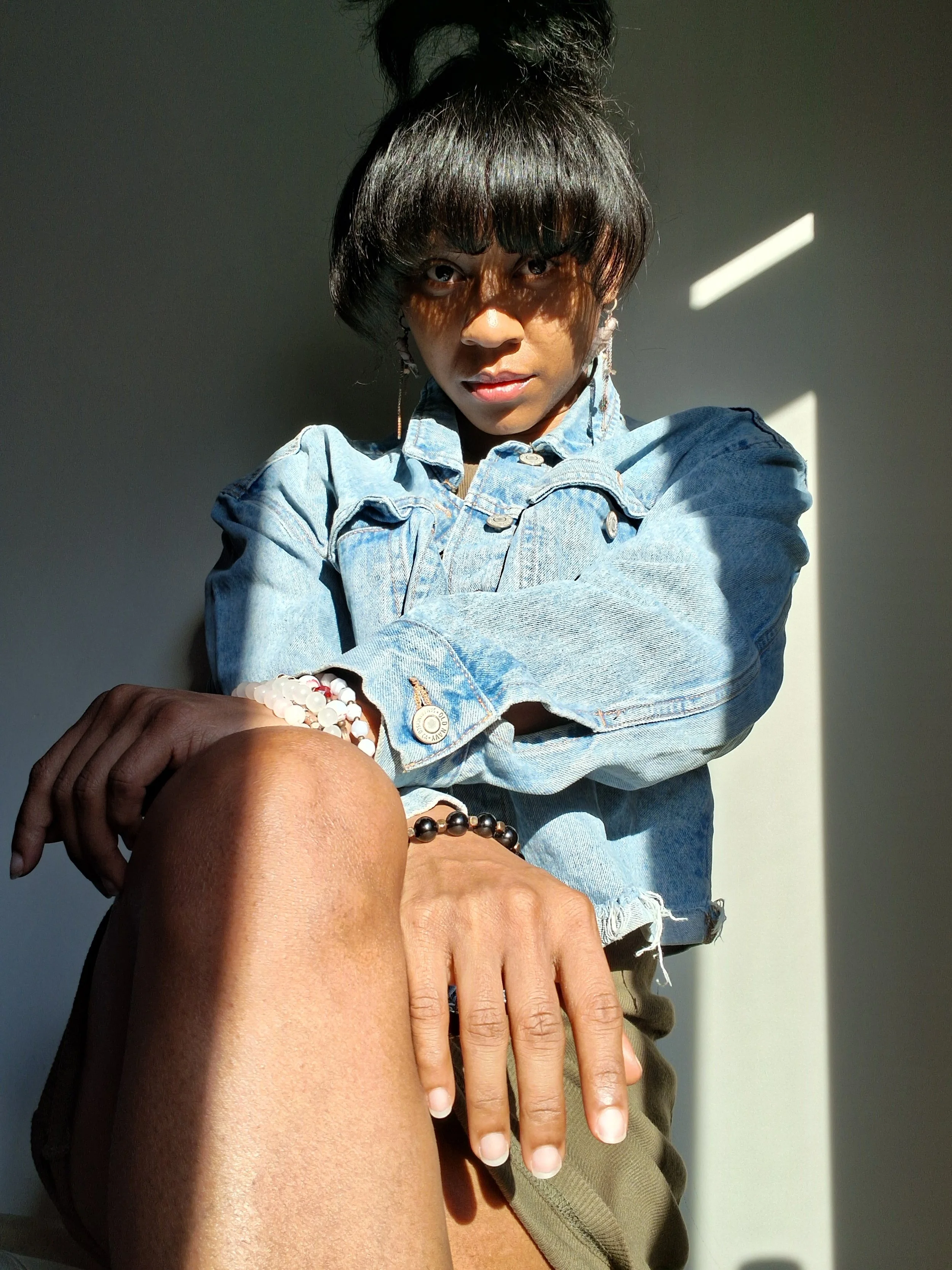 A person with dark hair tied up, wearing a denim jacket, and bracelets, sitting outside in sunlight with shadows casting on their face.