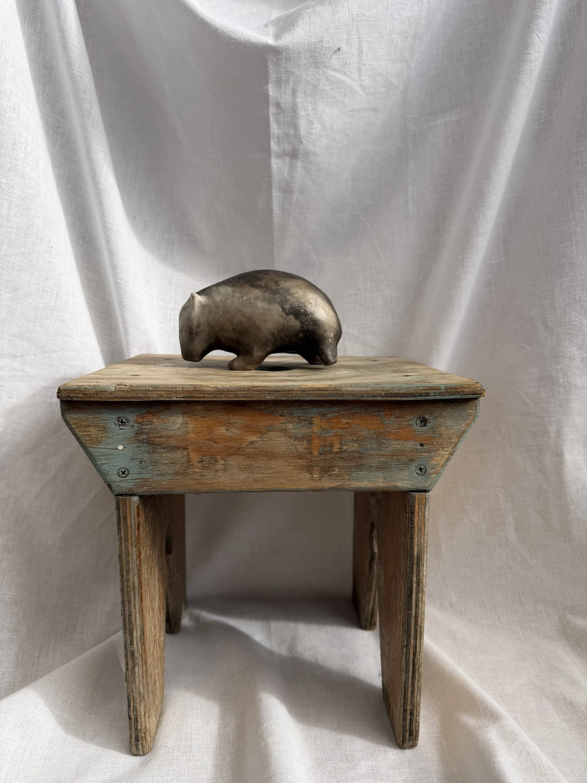 Large sized wombat sculpture, sitting on a wooden shelf with a white fabric backdrop.