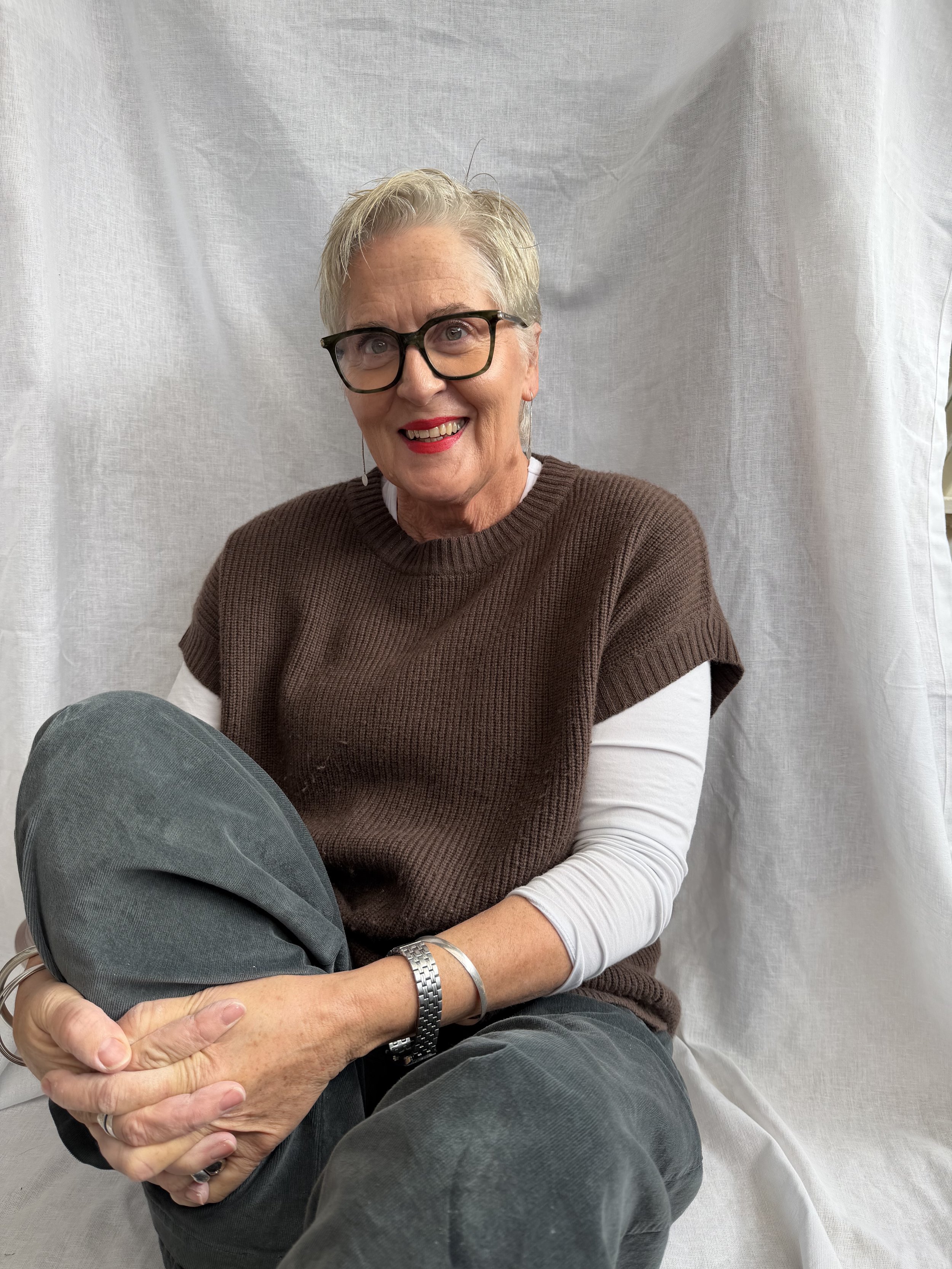 The artist, Fiona Barrett, smiling. Wearing a brown sweater with a white long-sleeve shirt underneath, sitting against a light-colored fabric backdrop.