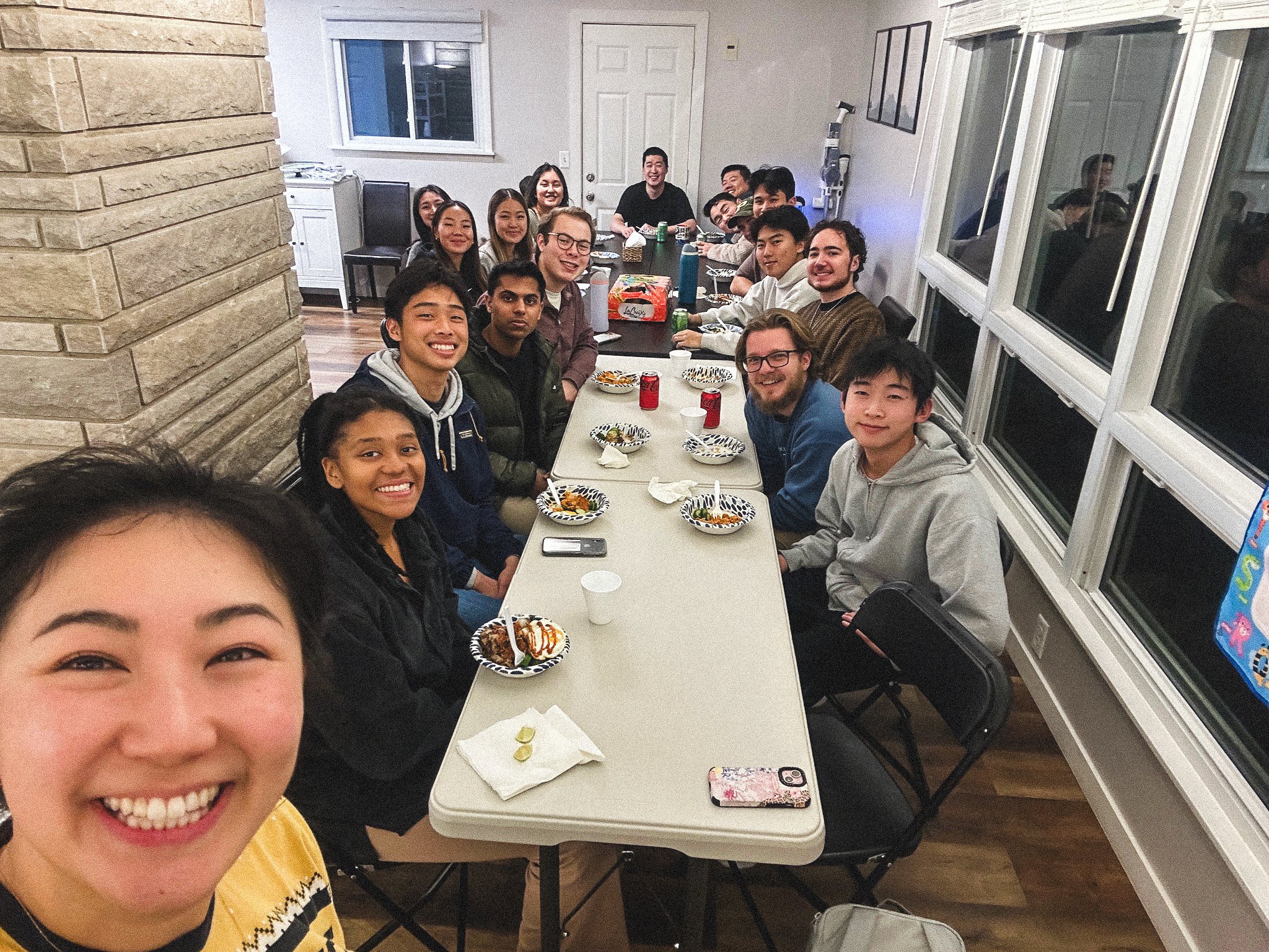 Group of people gathered around a long table at a celebration, smiling for the camera, with plates of food and drinks on the table.