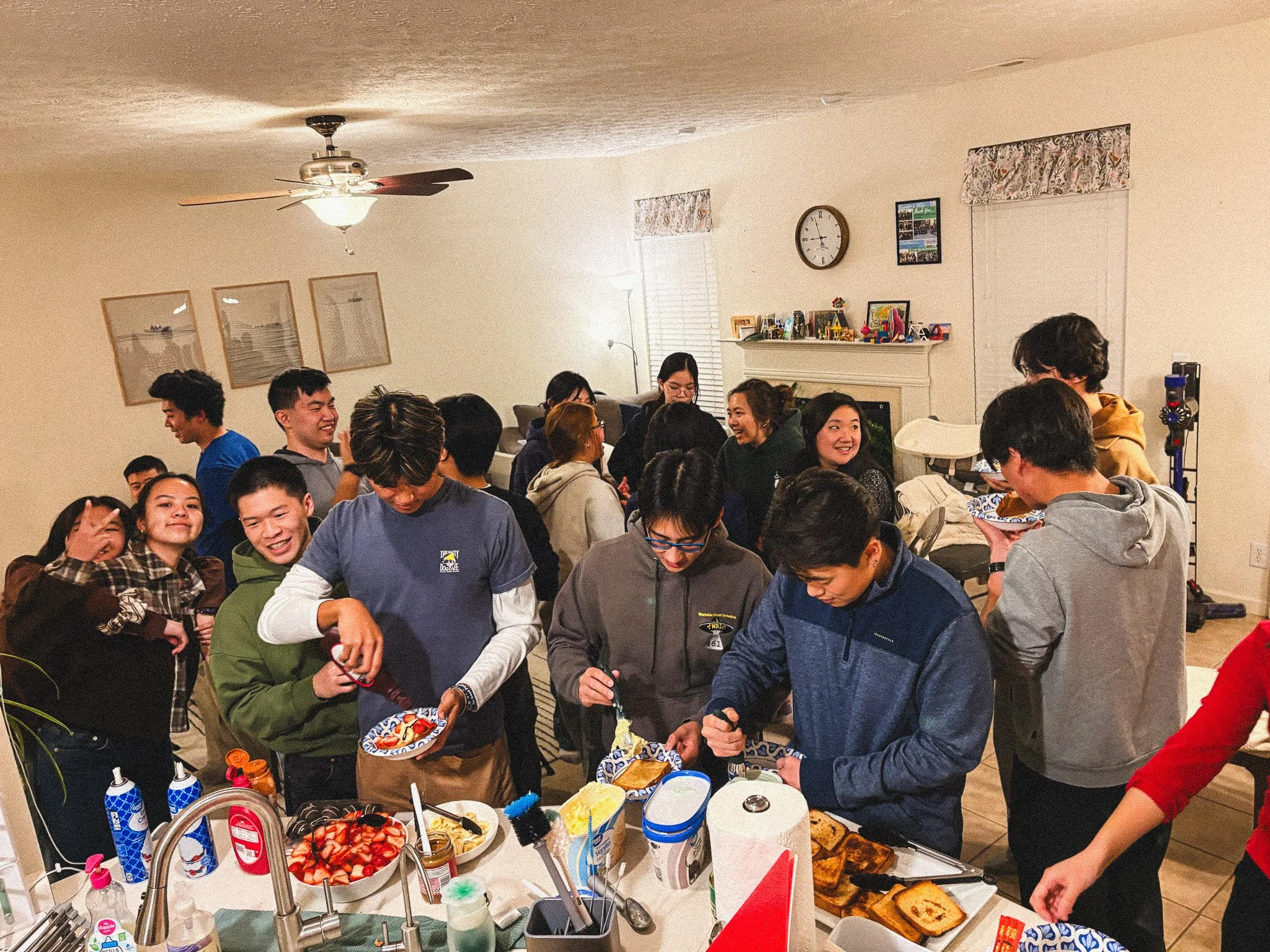 Group of diverse young adults gathering around a table filled with food, serving and chatting in a living room with a fireplace, wall clock, and framed pictures.