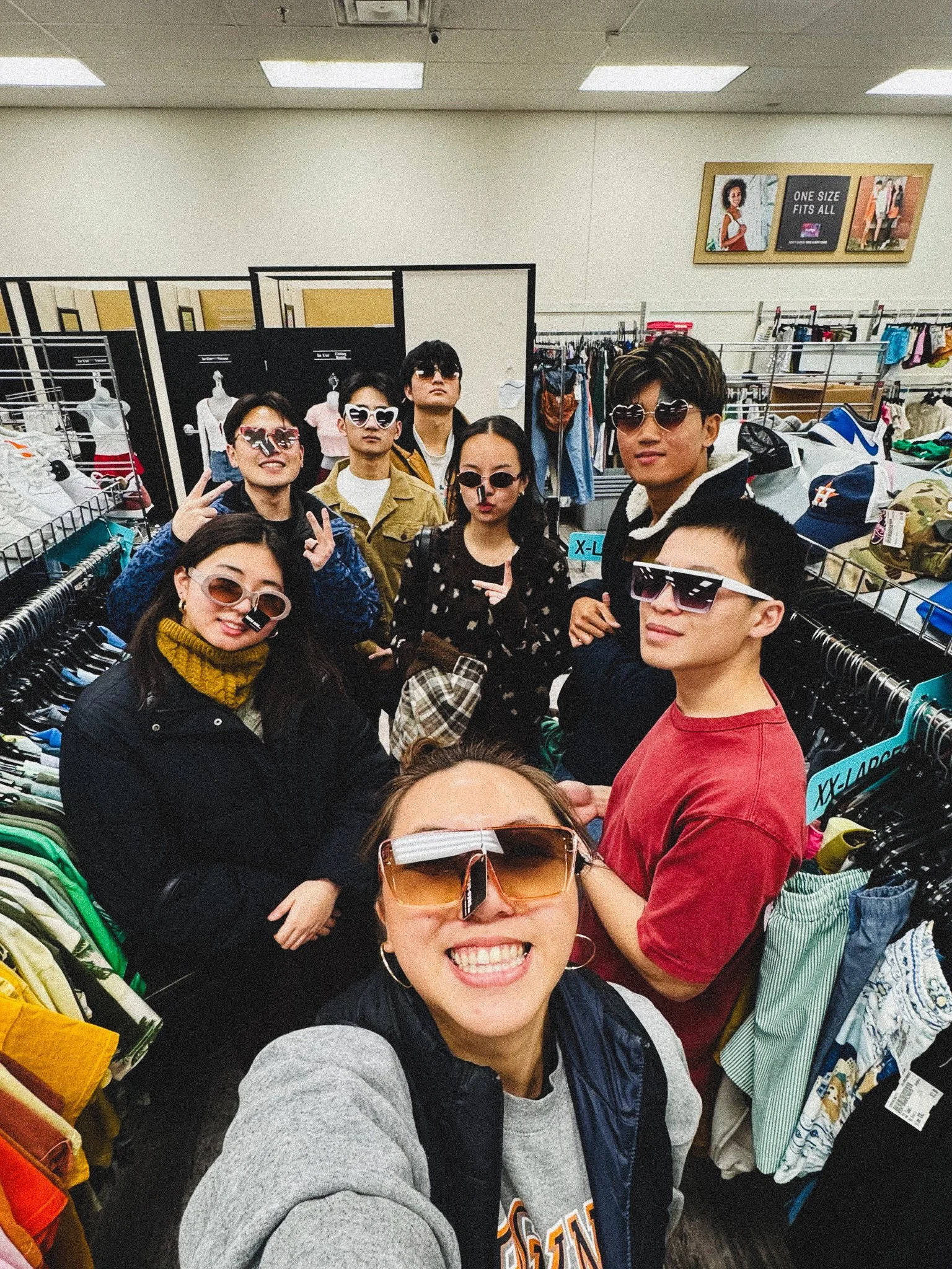 Group of seven young adults in a clothing store, taking a selfie together. They are all wearing sunglasses and smiling.