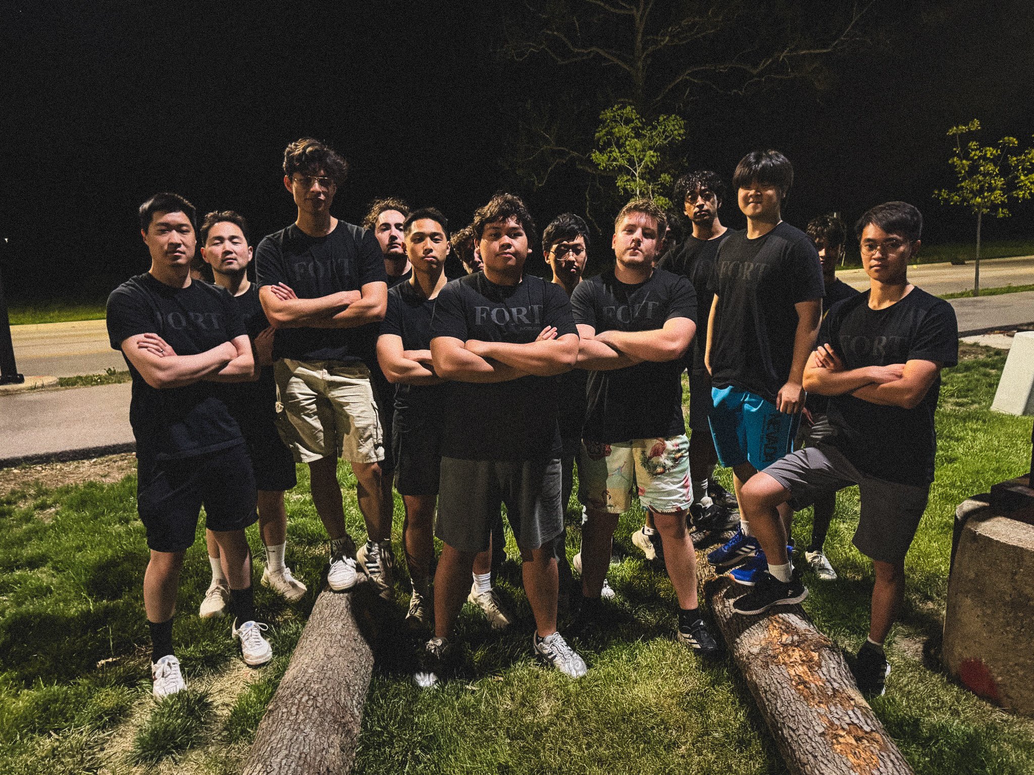 A group of young men standing outdoors at night, some crossing their arms and others with their hands at their sides, posing under a large fallen tree with a grassy area and trees in the background.