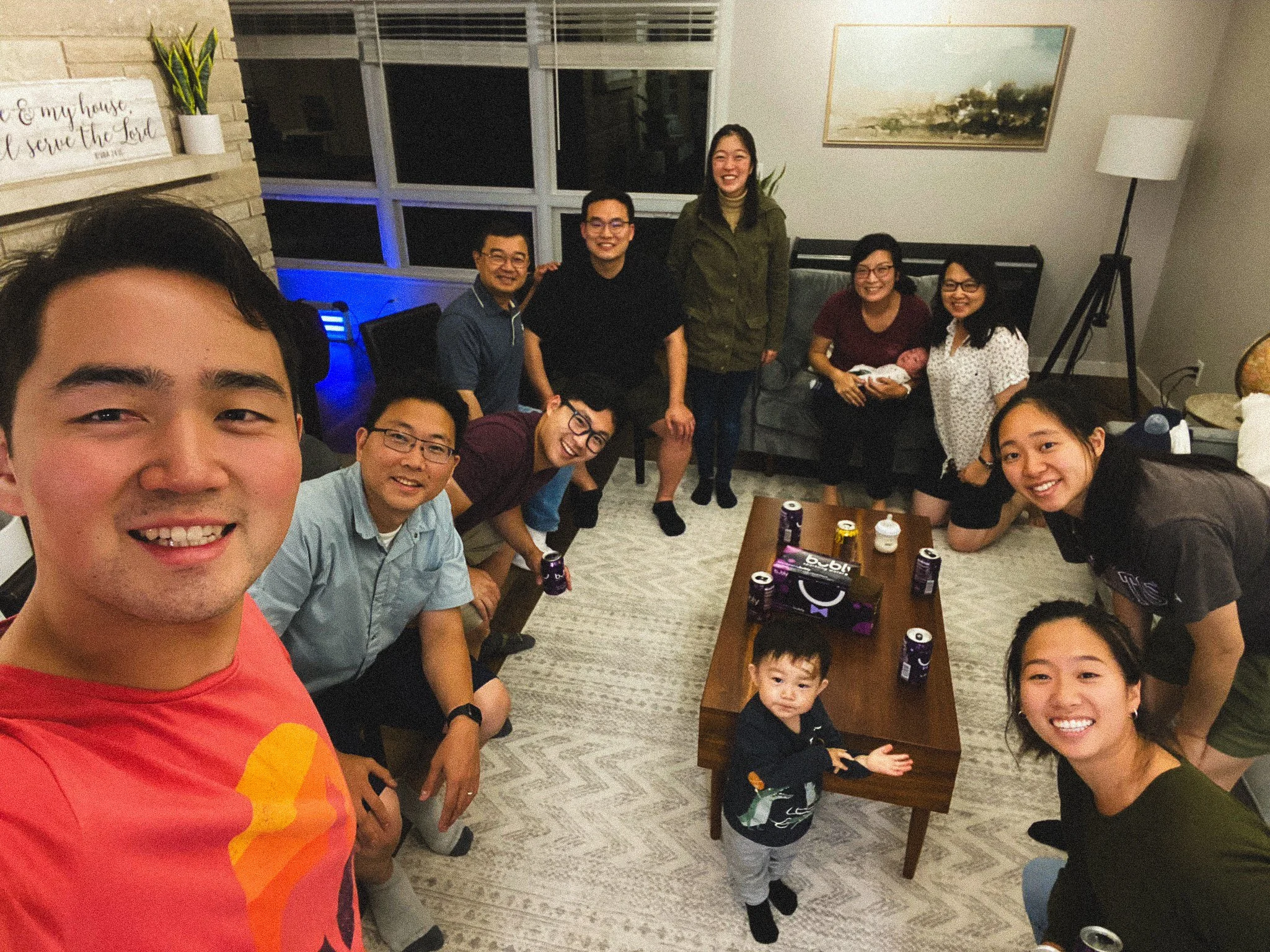 A group of people, including adults and children, gathered indoors for a celebration, with some smiling and posing for a photo. There is a woman holding a newborn baby, and several cans of soda on a coffee table.
