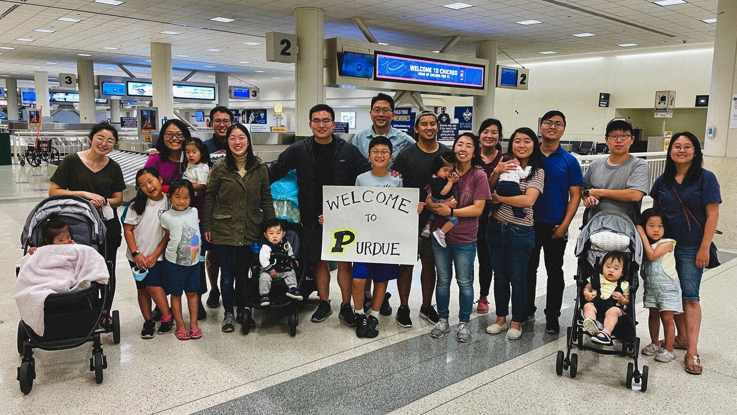 Group of people at an airport holding a sign that says 'Welcome to Purdue'. There are children and adults, some with strollers, in a spacious terminal with several departure screens overhead.