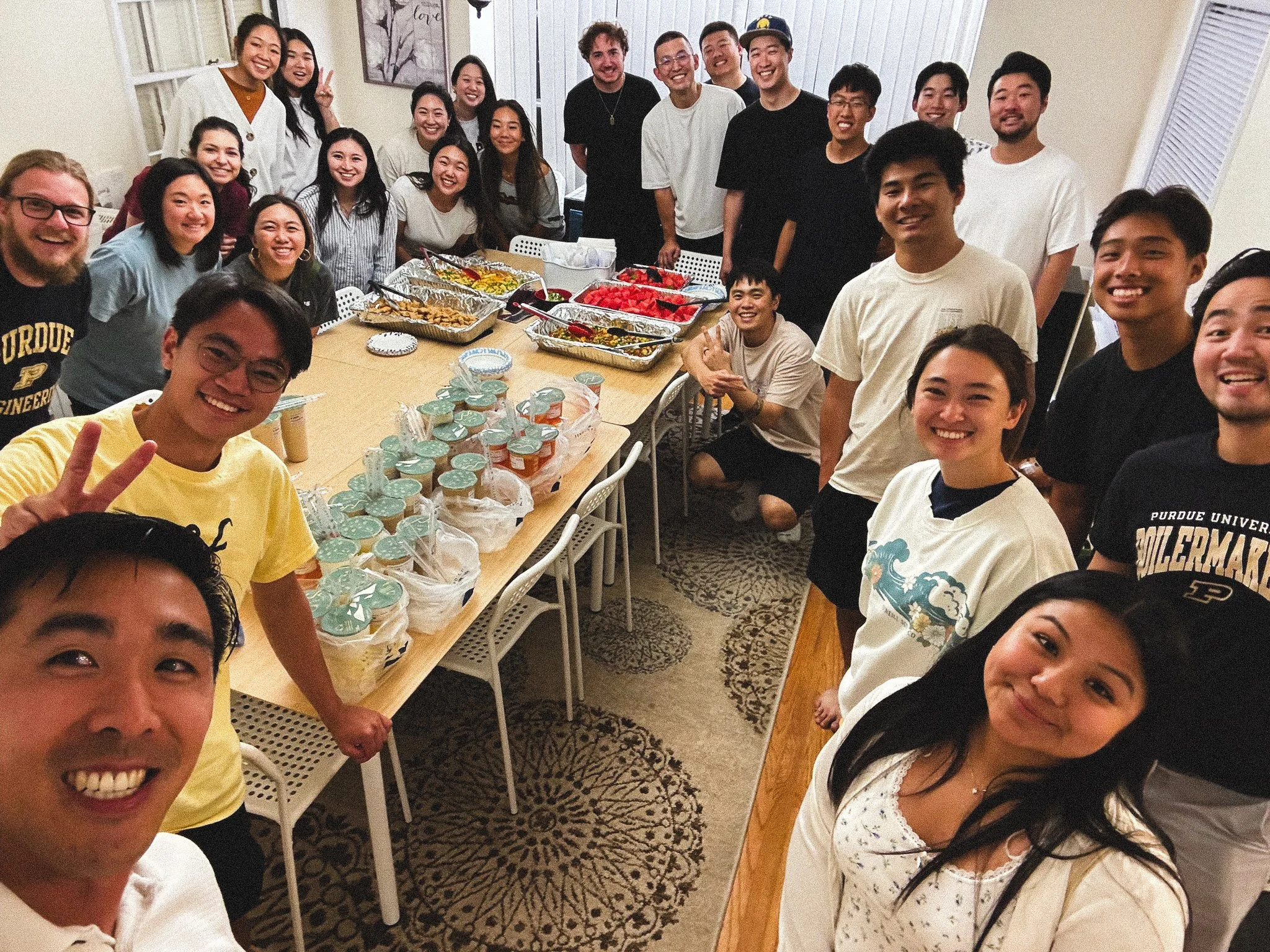 Group of young adults smiling and gathered around a table with food and drinks, indoors.