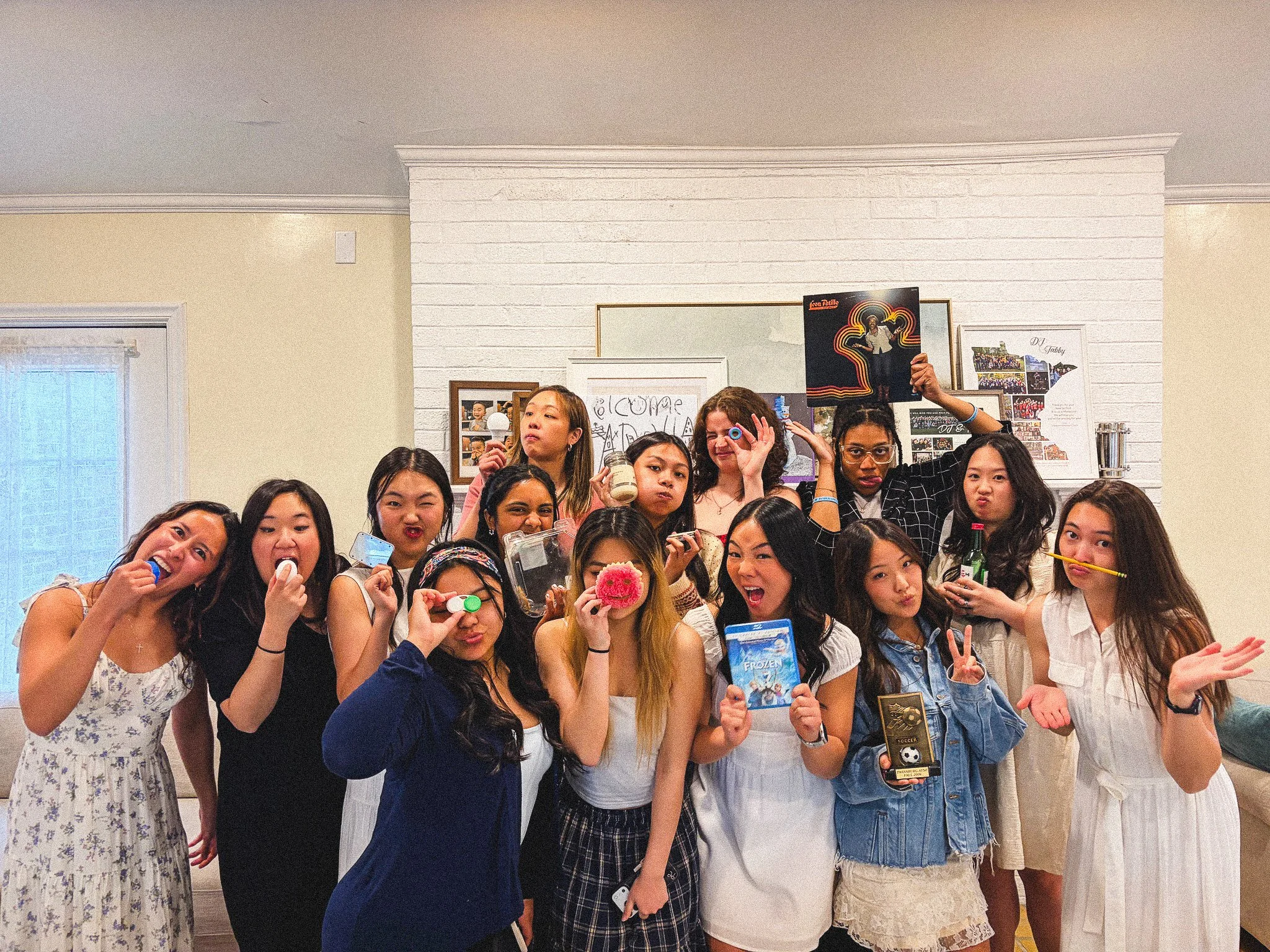 Group of young women posing together at a social gathering, some holding drinks, snacks, or objects, standing in front of a white brick wall decorated with photos and posters.