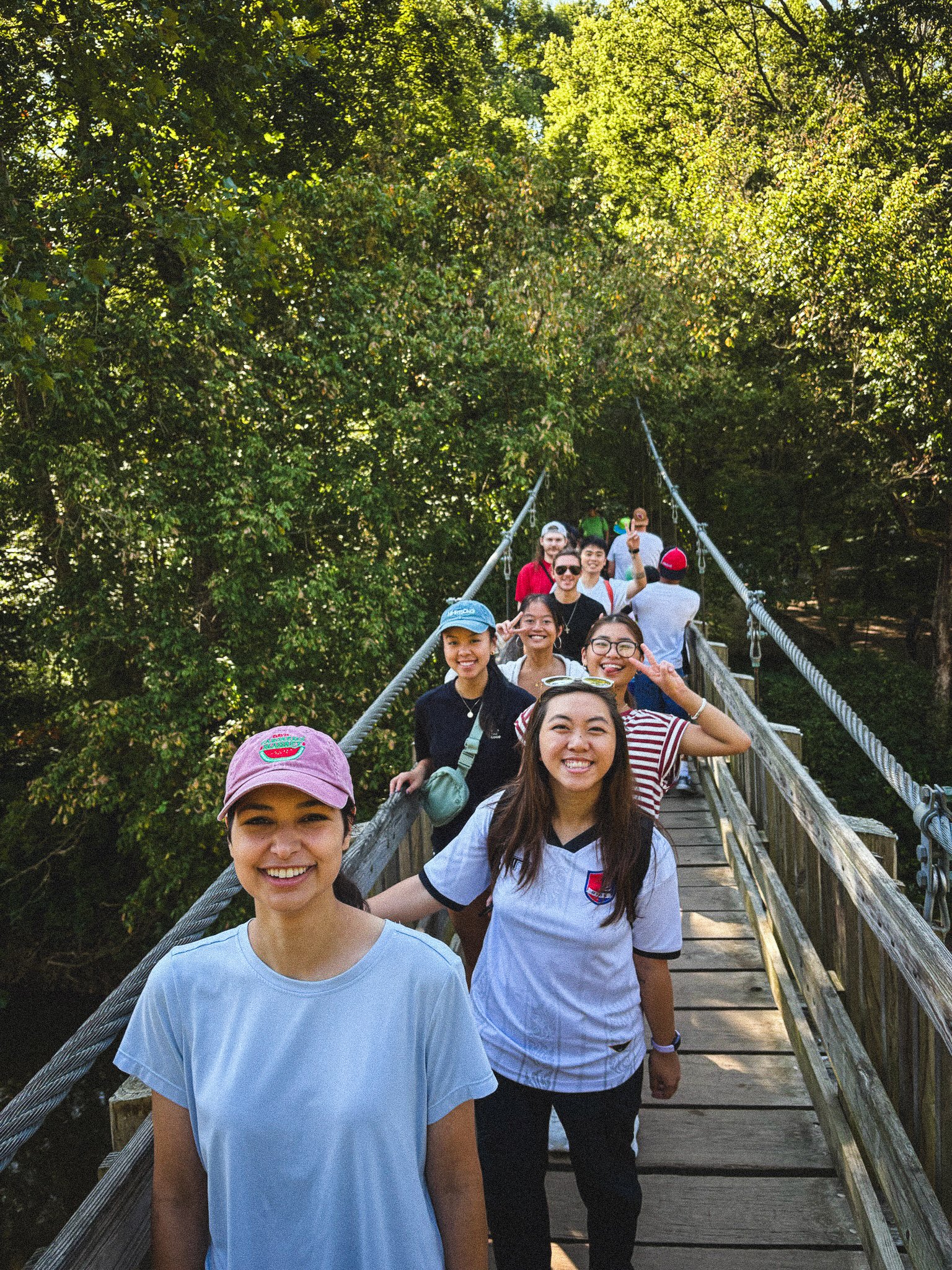 A group of people walking across a wooden suspension bridge in a forest, smiling and making peace signs.