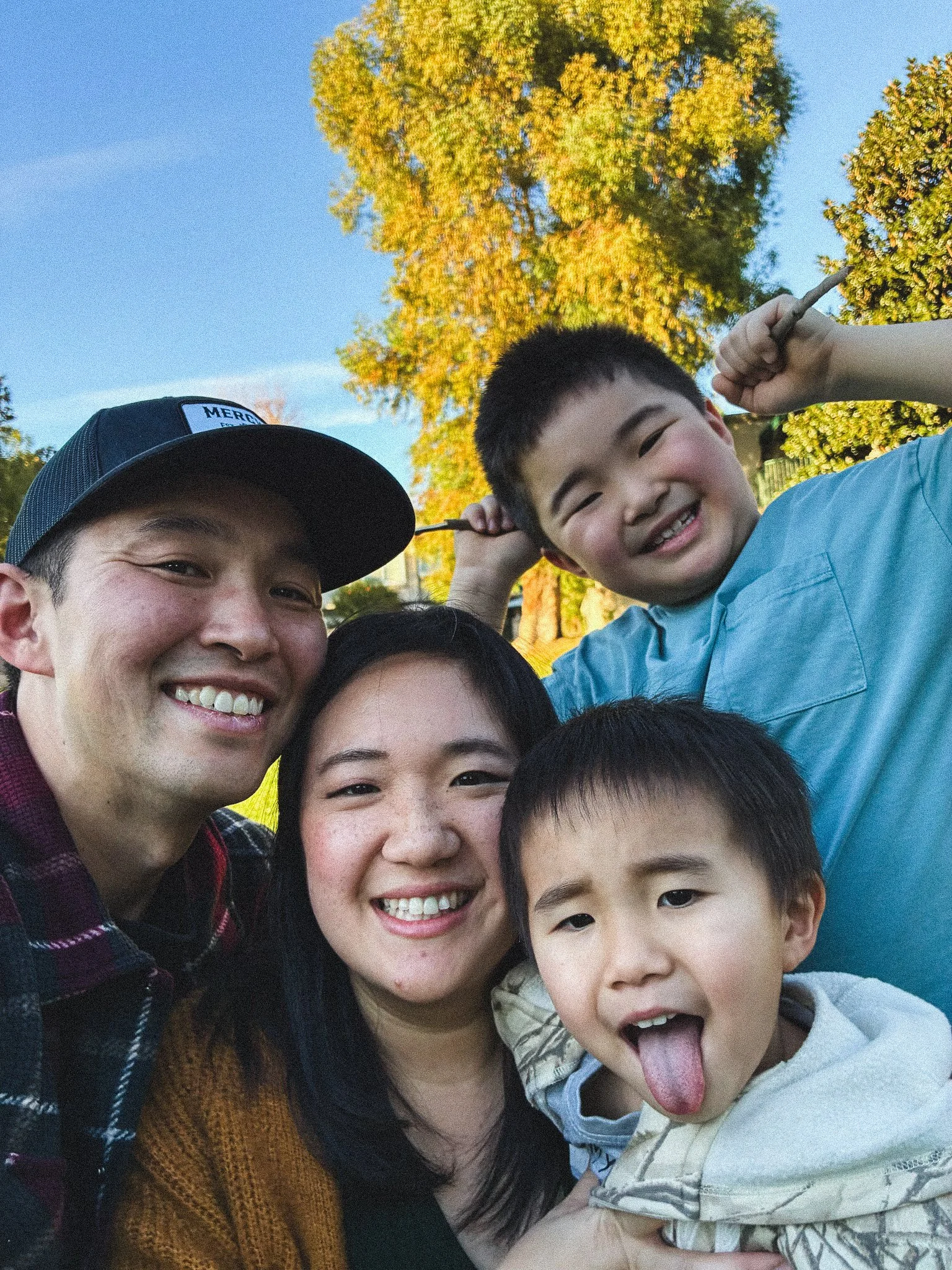 A group of smiling children and young adults taking a selfie outdoors, with trees and a clear sky in the background.
