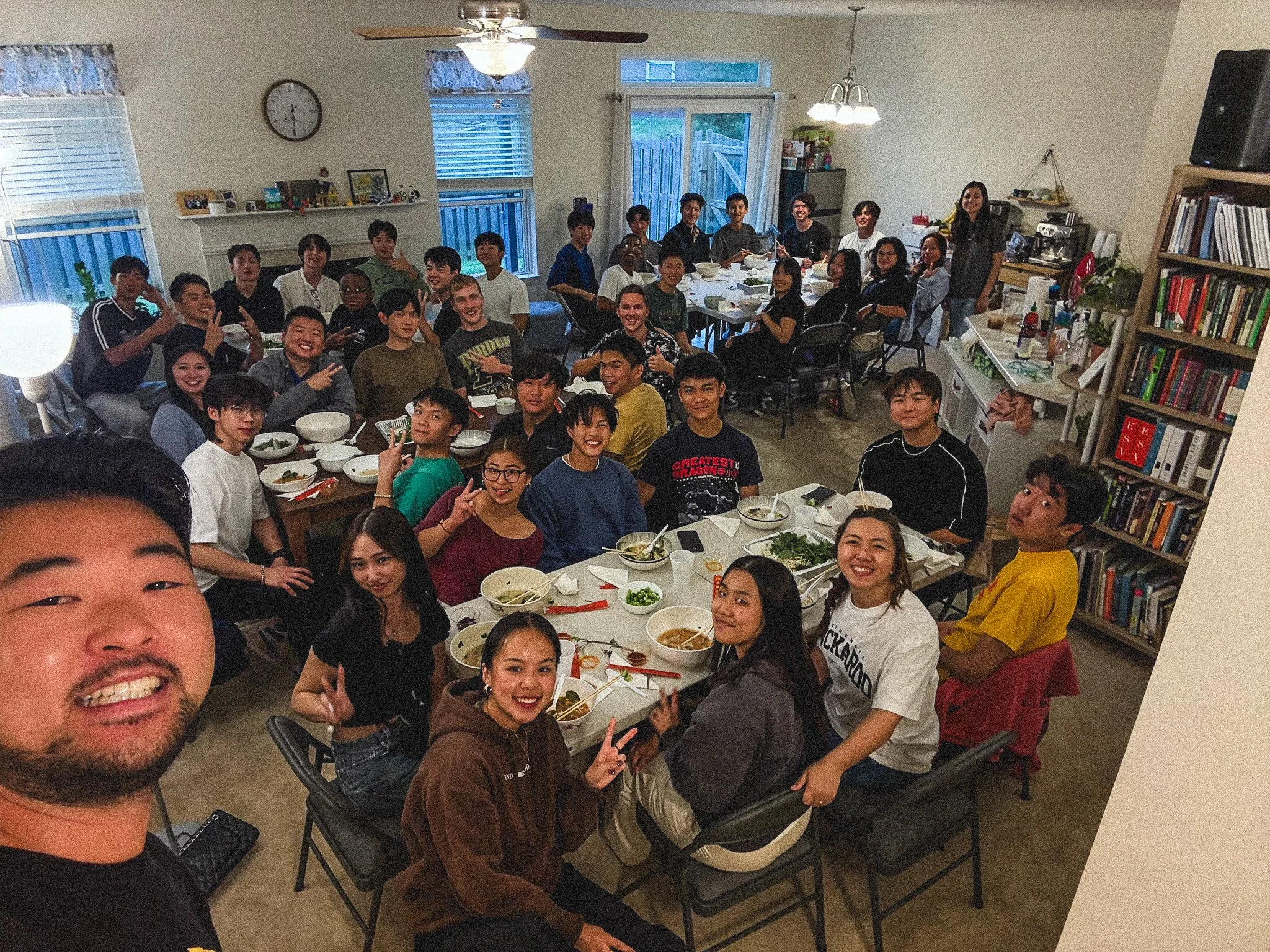 Group of people gathered around a dining table with food, smiling and making peace signs for a photo in a living room with bookshelves, windows, and a door leading outside.