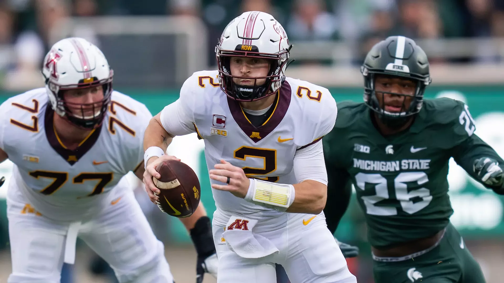 A college football game with a player from Minnesota wearing a white jersey and a helmet, running with the football while being pursued by players from Michigan State in green jerseys.