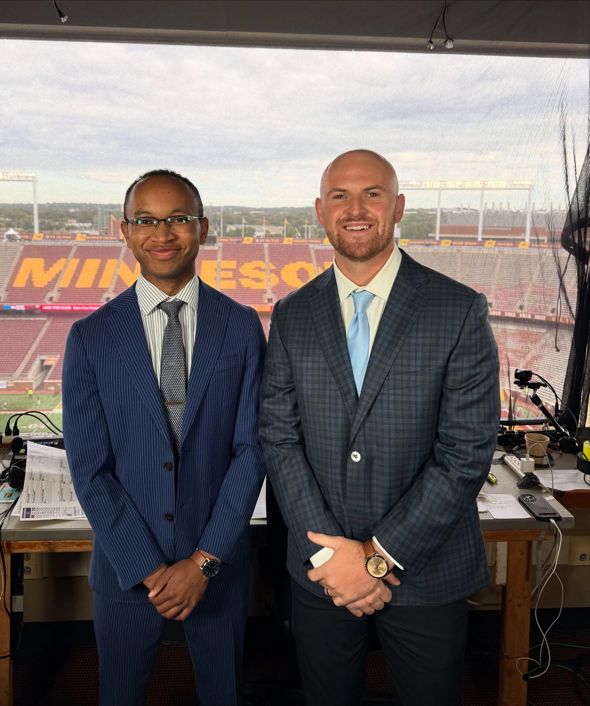 Two men dressed in suits standing in a press box at a football stadium, with empty stadium seats and field visible through the window behind them.