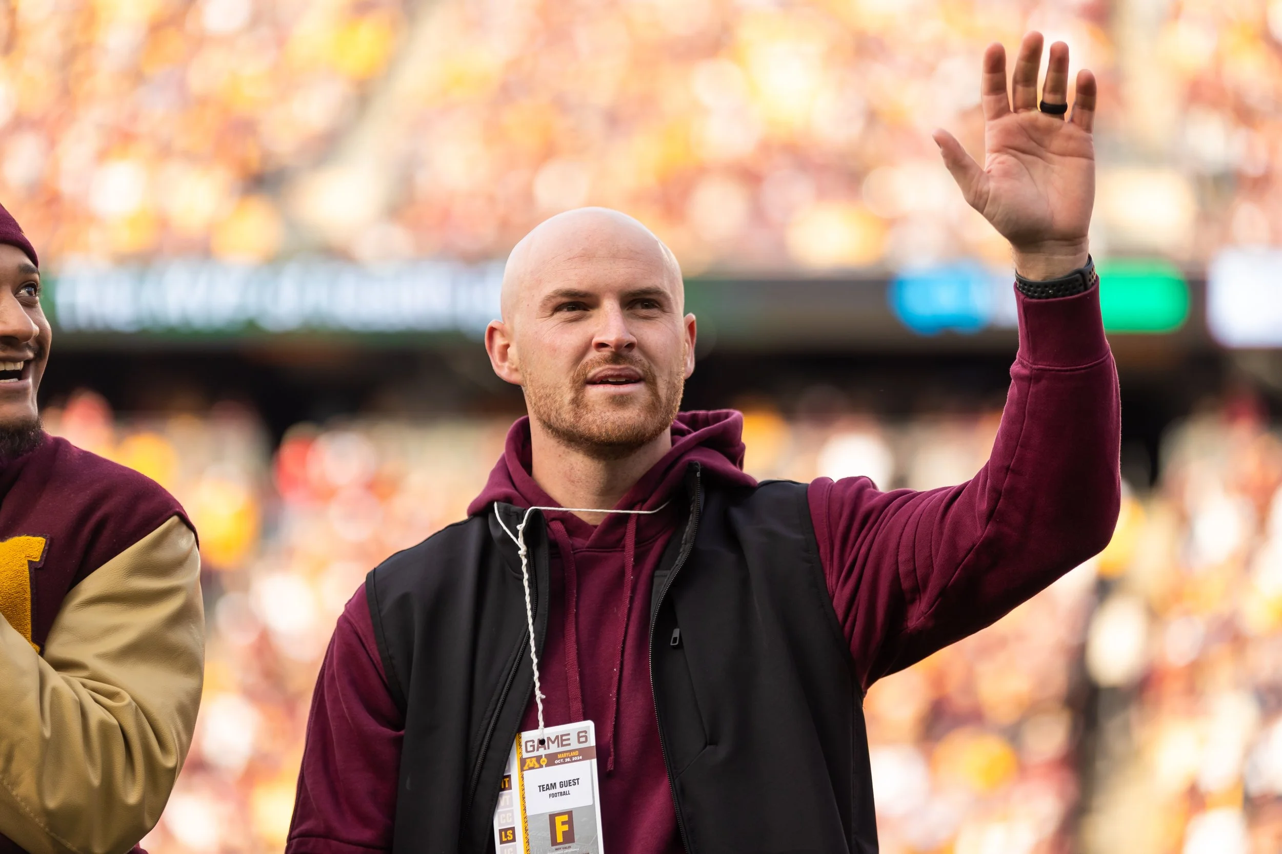 A man with a shaved head and beard raising his hand in a greeting gesture at a public event, with blurred trees in the background and a person wearing maroon clothing partially visible on the left.