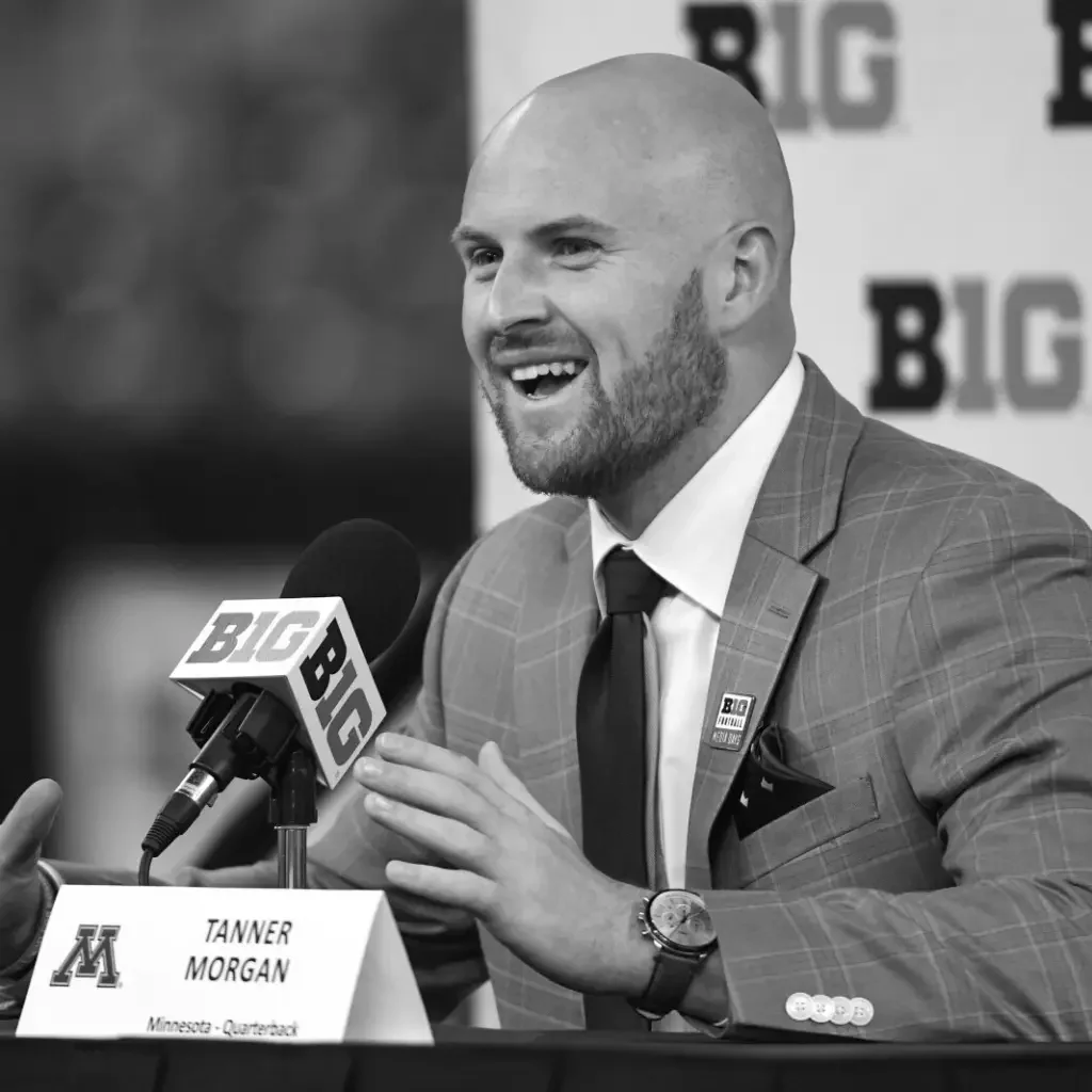 A man, likely Tanner Morgan, wearing a checkered suit jacket, a white shirt, and a dark tie, speaking into a microphone at a press conference. He has a bald head, a beard, and is smiling. A nameplate in front of him indicates his name, profession as quarterback for Minnesota, and the event appears to be related to Big Ten sports.