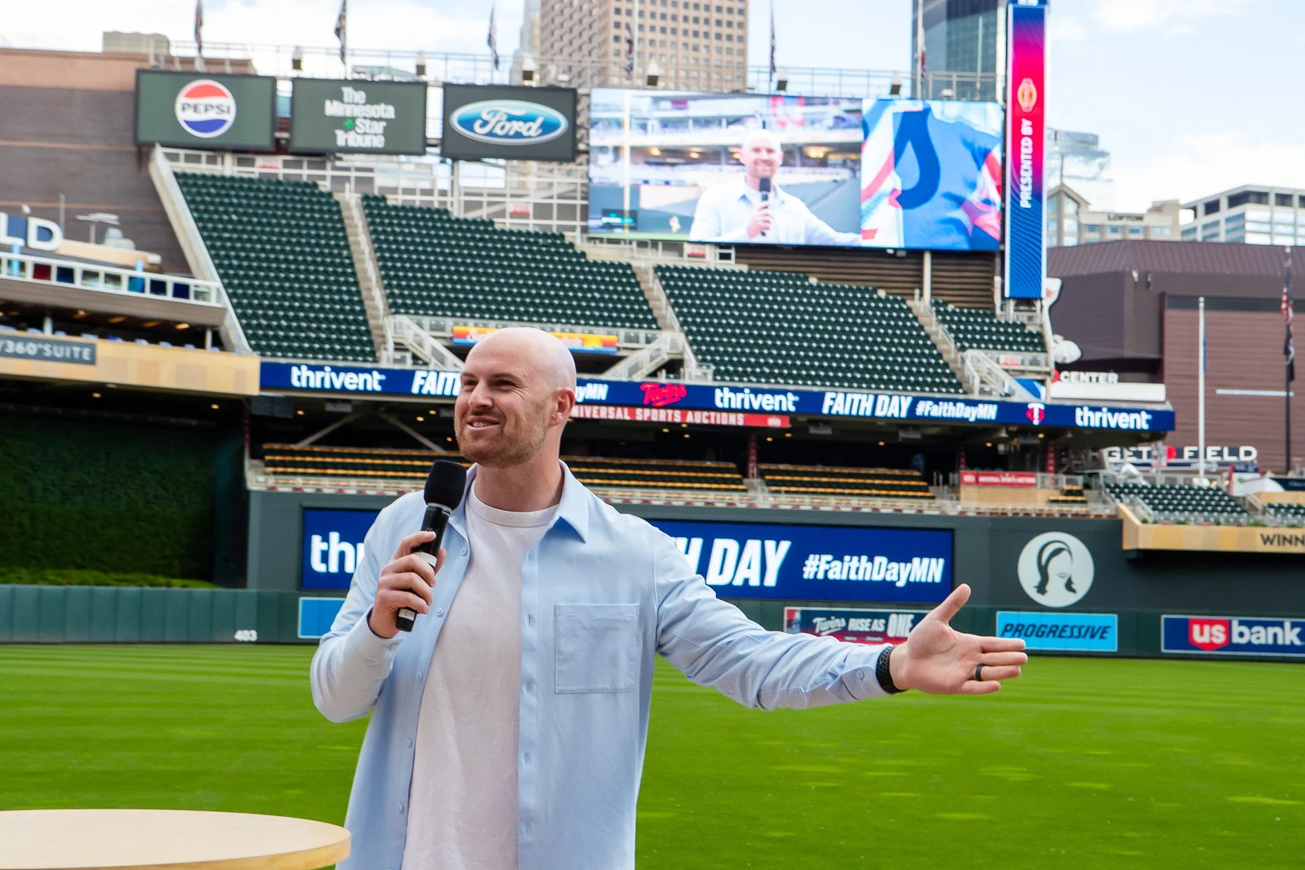 A man in a light blue shirt and white t-shirt holding a microphone and speaking at a baseball stadium, with empty green stadium seats, electronic advertisement banners, and a large screen showing him in the background.