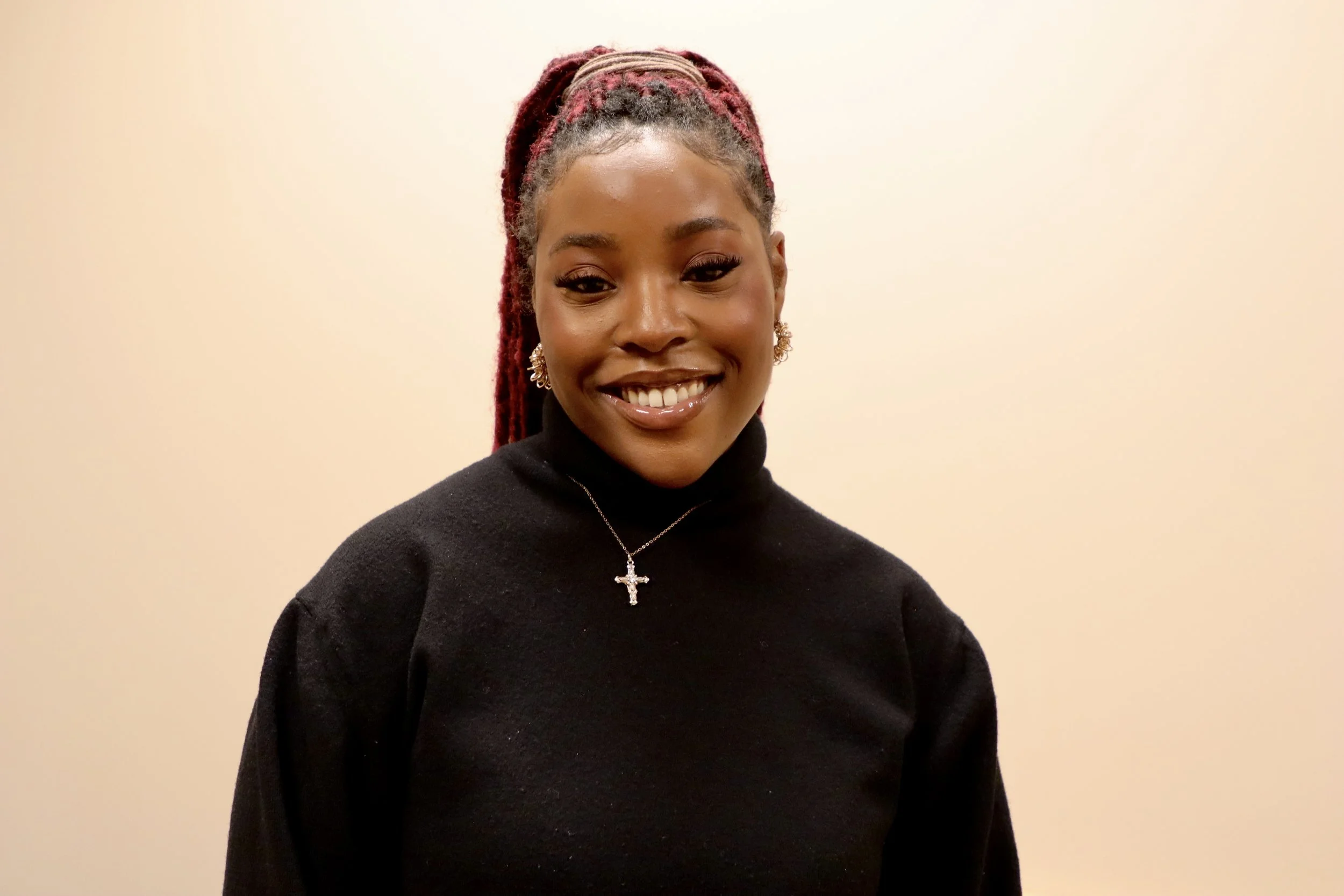 A woman with dreadlocks in a red hue, smiling and wearing a black top, a cross necklace, and earrings, standing against a plain light-colored background.