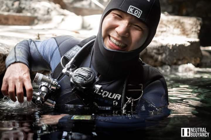 Swim instructor and technical diver Andy, is wearing a full scuba gear with a hood, regulator and wetsuit, standing chest deep in a cenote, smiling at the camera