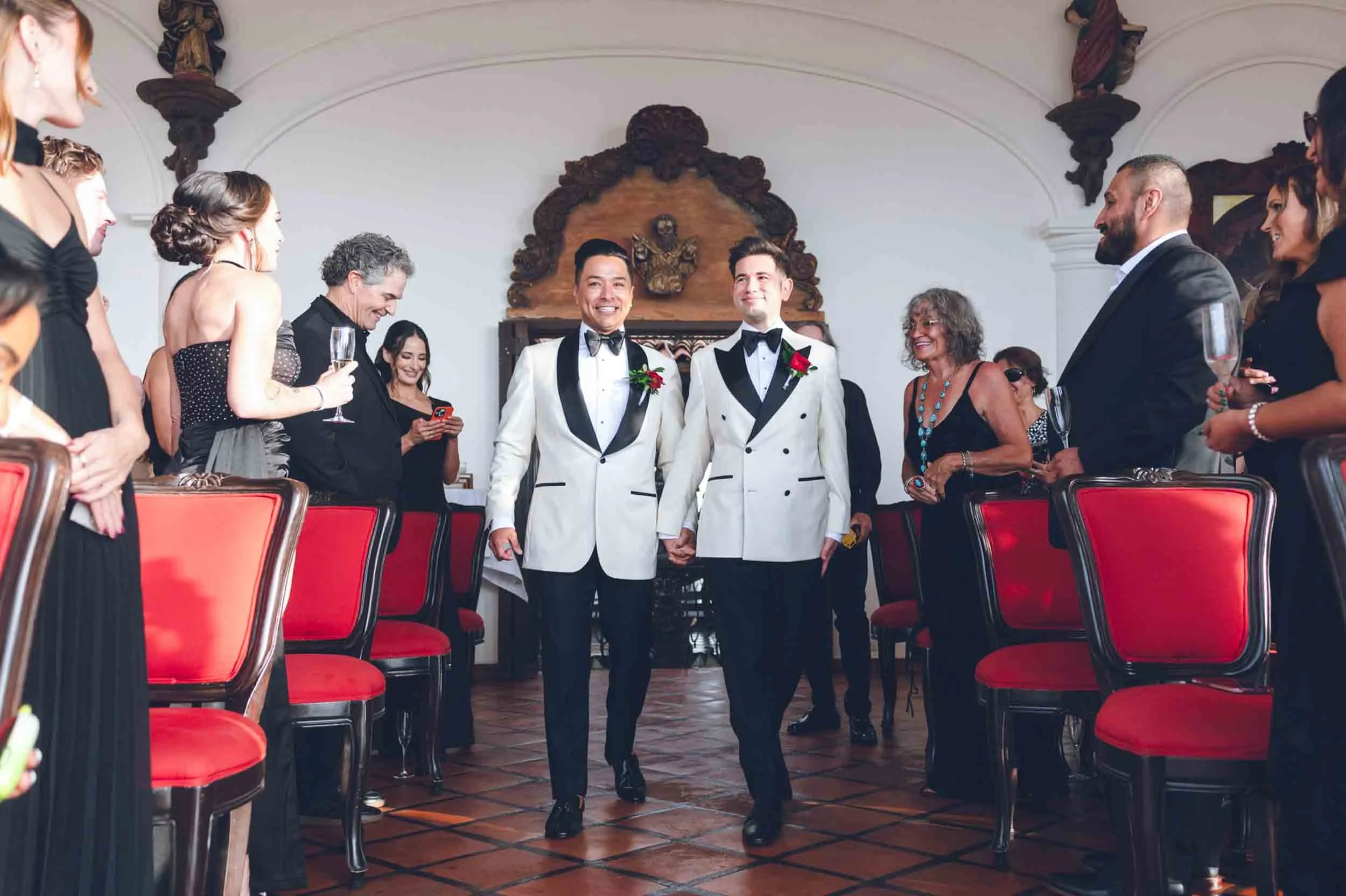 Entrance of gay couple holding hands walking down the aisle at their wedding ceremony, surrounded by seated guests in formal attire.