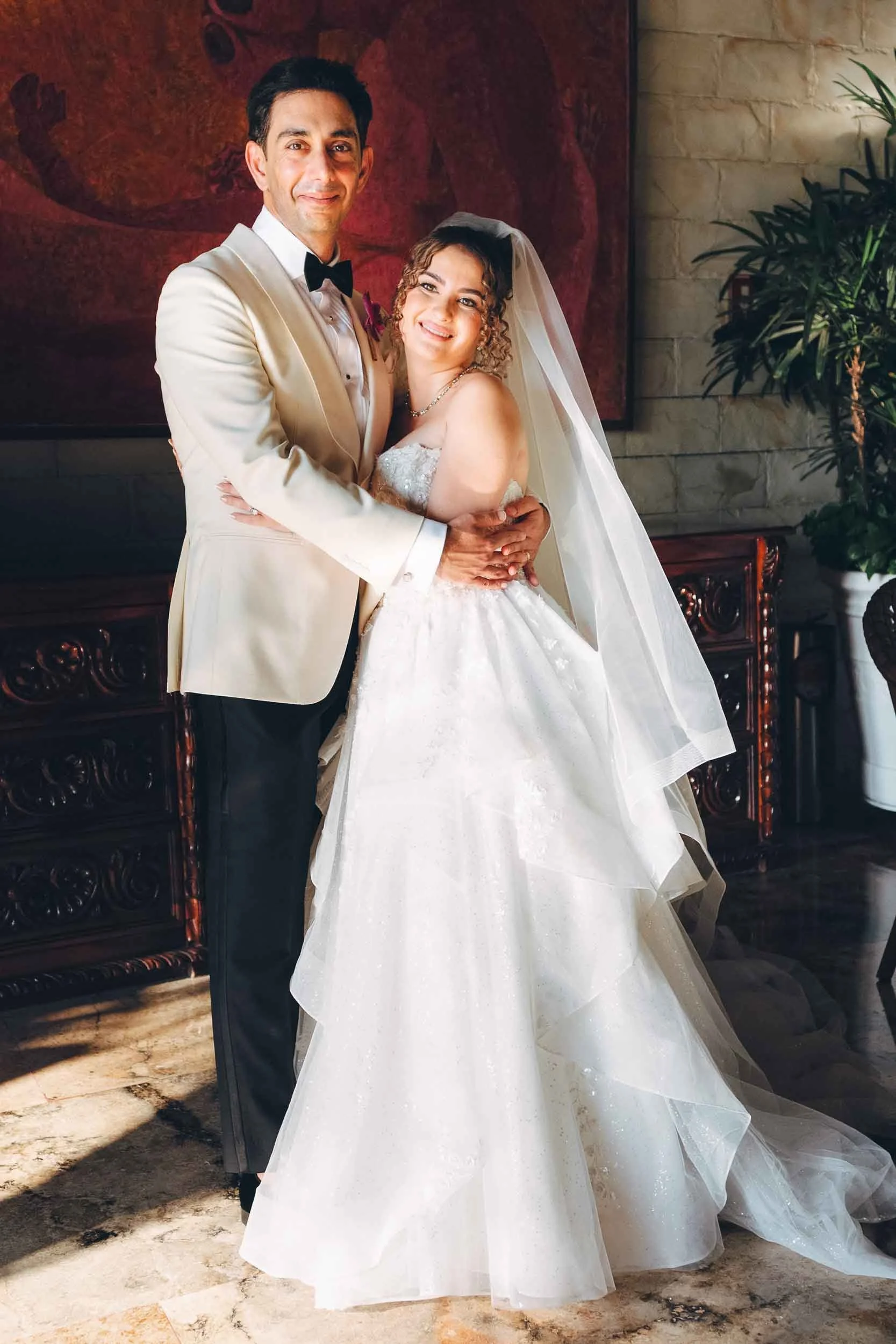 A couple on their wedding day, dressed in formal wedding attire, standing indoors with a decorative backdrop and potted plants.