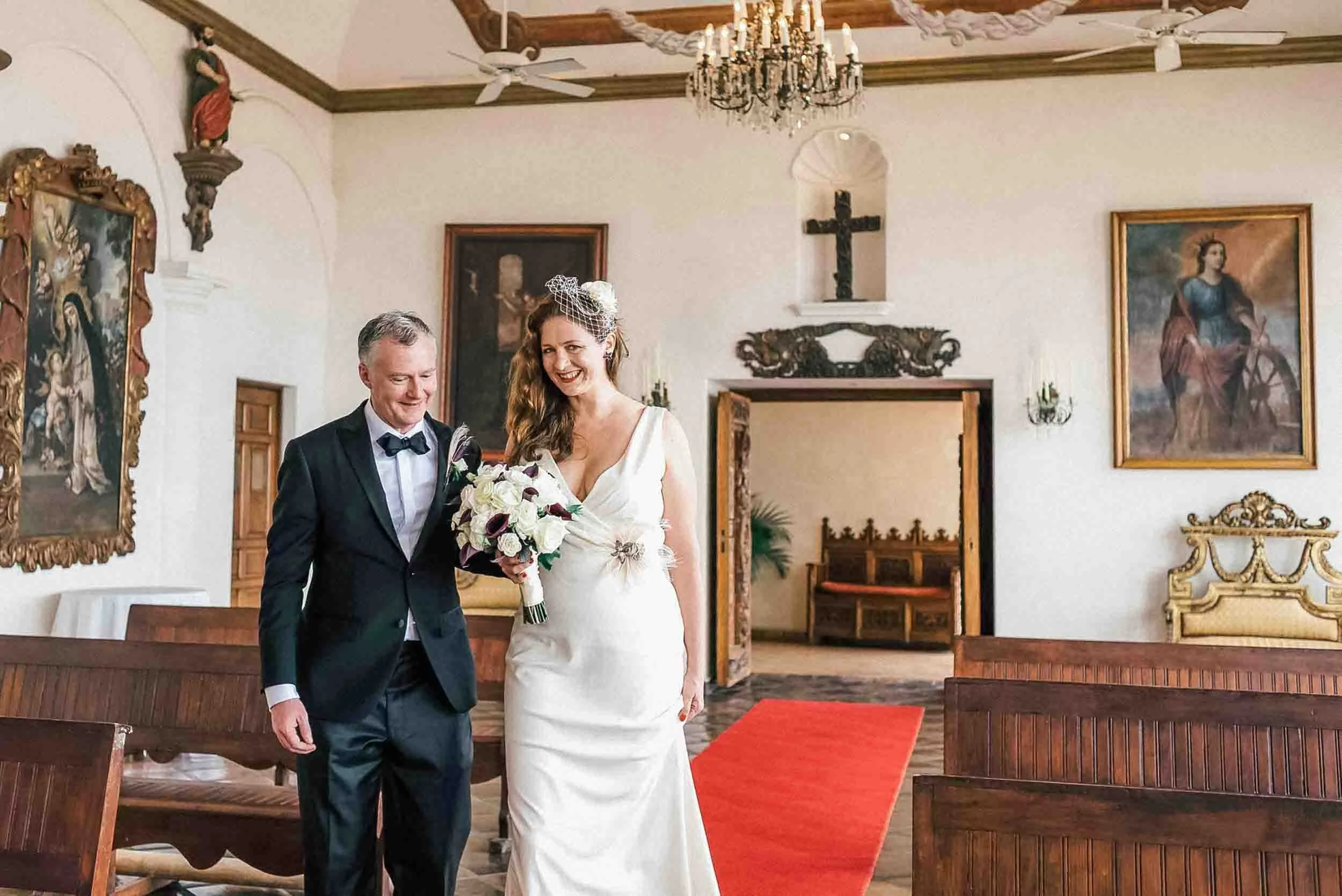 A bride and groom standing together. The bride is holding a bouquet of white flowers, and both are smiling. The venue has religious art on the walls, chandeliers, and wooden pews.