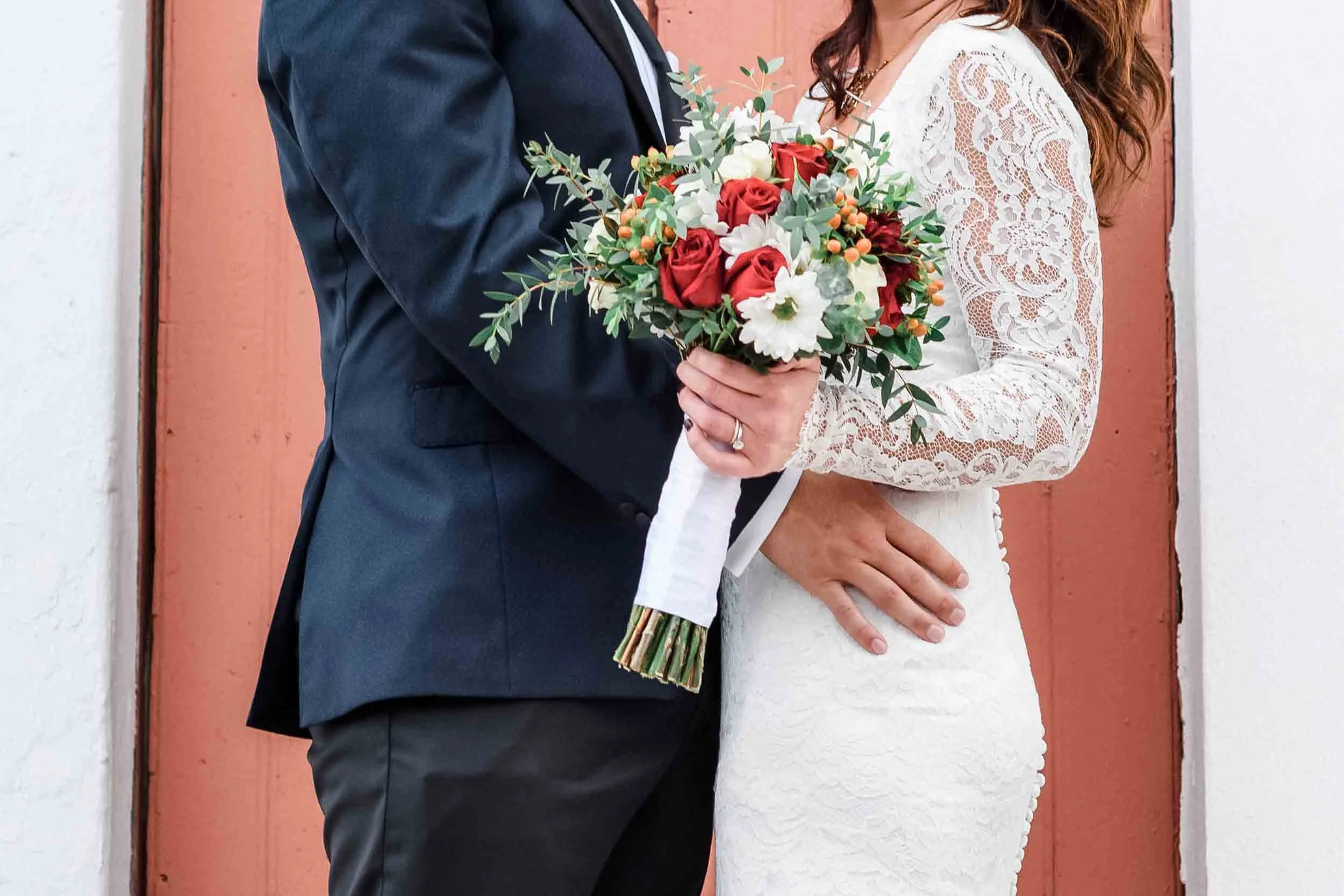 Close-up of a couple dressed in wedding attire, holding a bouquet of red, white, and green flowers in front of a wooden background.