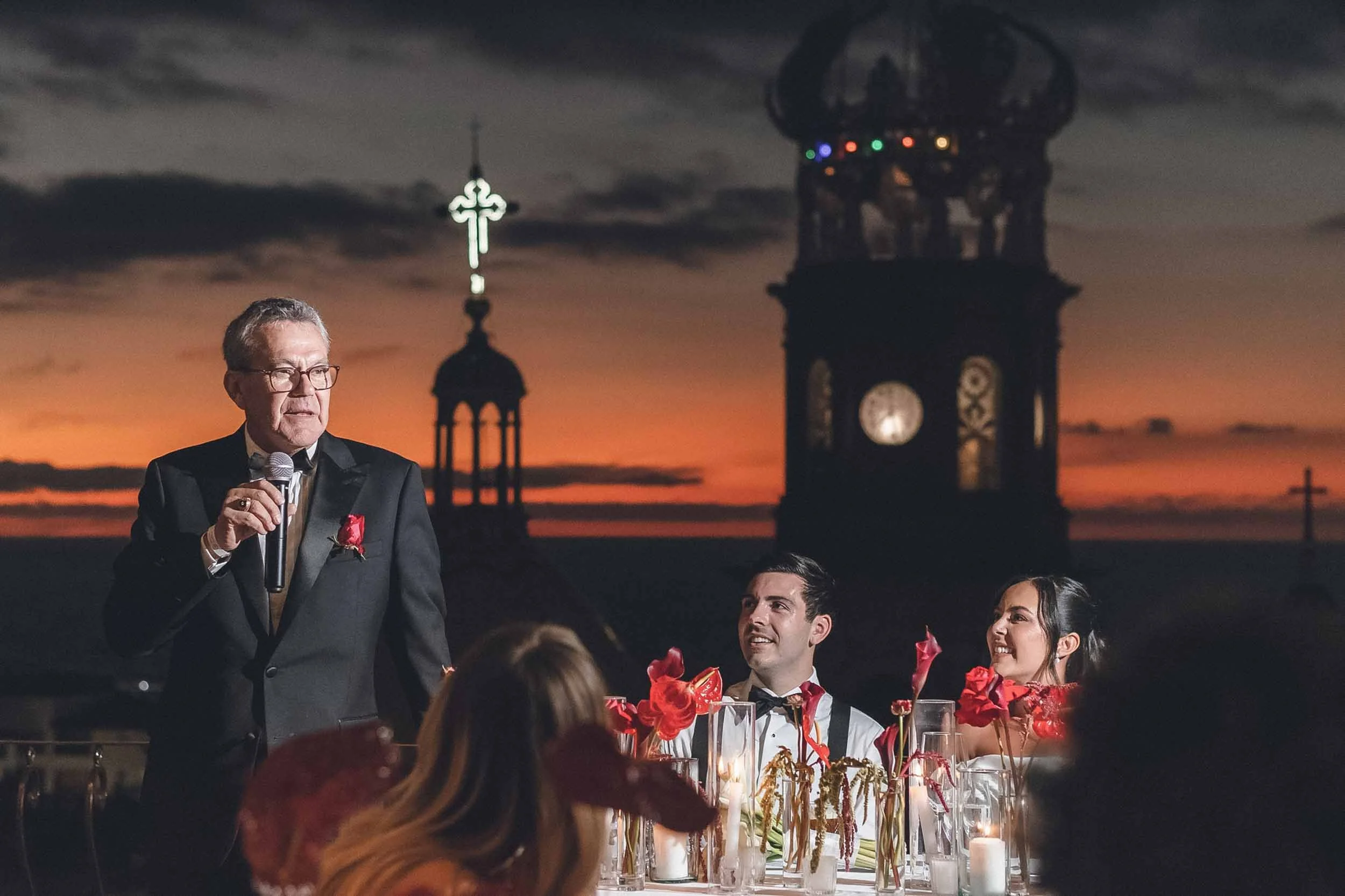 Father of the bride speaks at wedding reception during sunset, with a clock tower and a cross in the background, and seated guests enjoying the occasion.