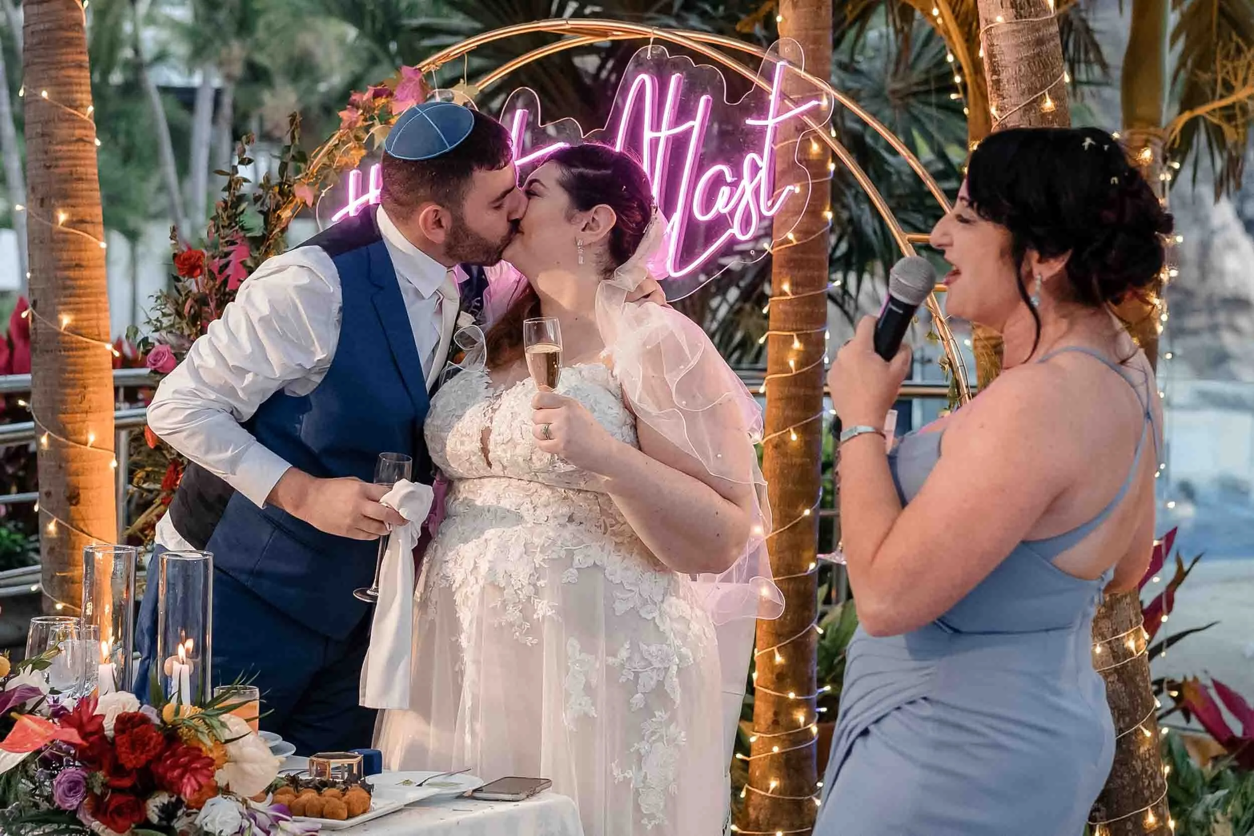 Bride and groom kissing during their wedding reception. The maid of honor holding a microphone is speaking nearby. There are decorated trees with string lights and a table with flowers and food in the foreground.