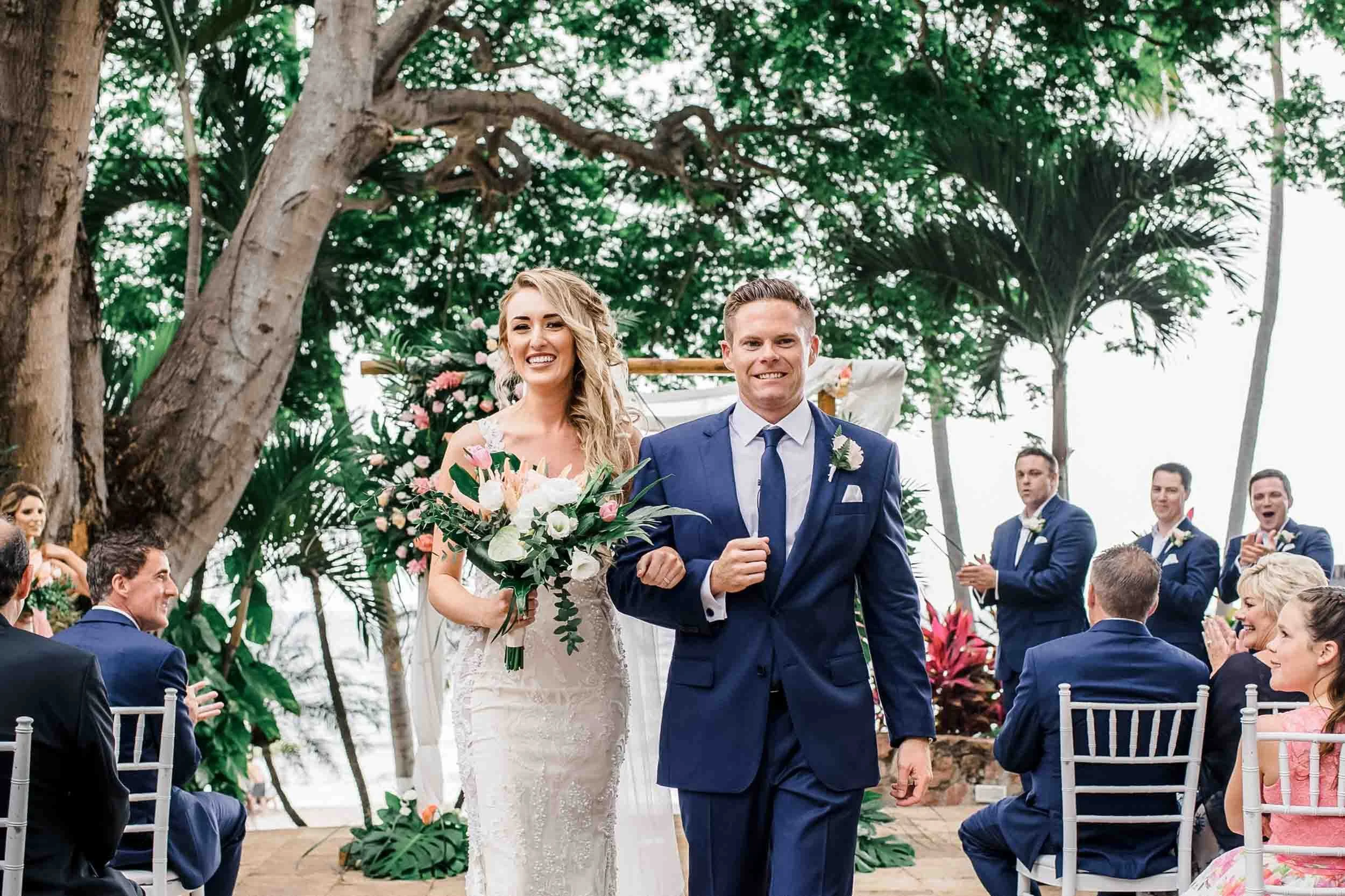 Bride and groom walk down the aisle at outdoor wedding ceremony at the Hyatt Ziva. Guests are seated and standing around them, clapping and smiling.