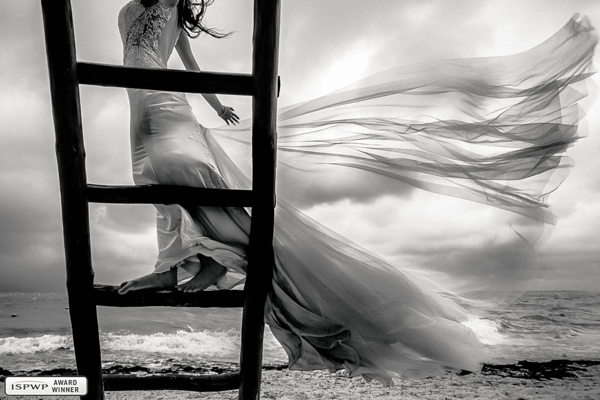 Bride on ladder while strong wind makes her dress fly with an ocean in the background.
