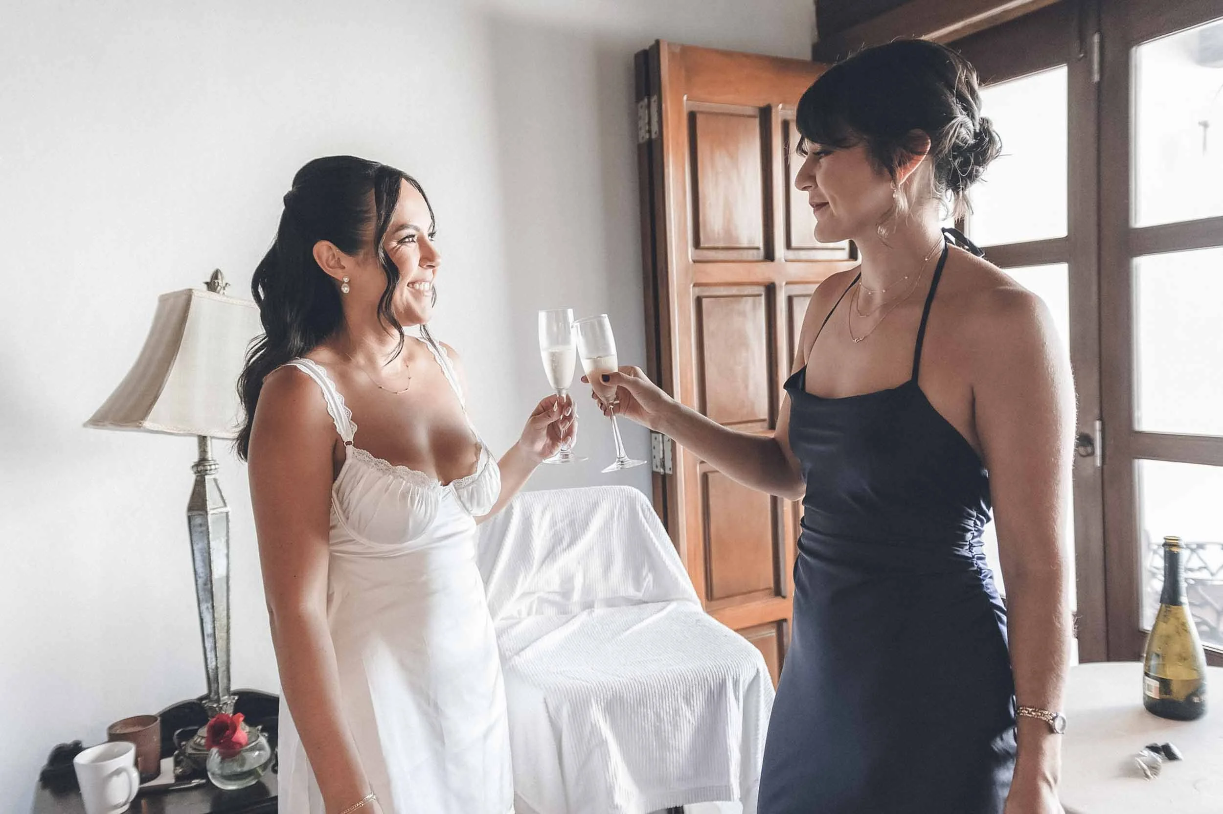  Bride and sister smiling at each other and clinking champagne glasses. 