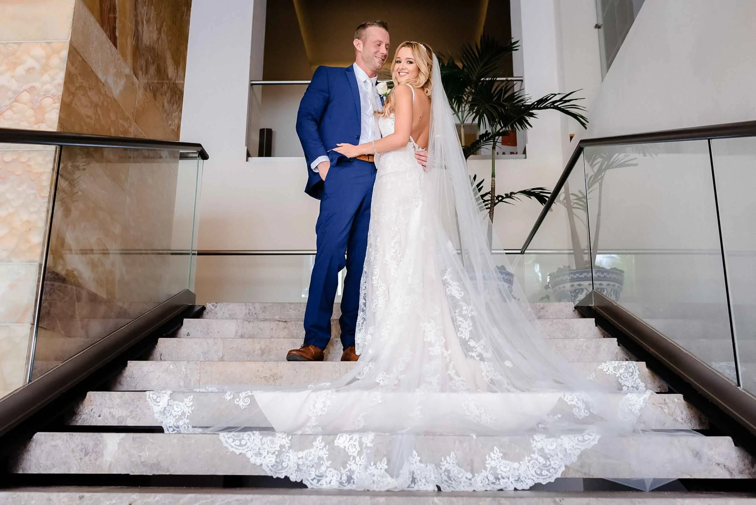 A bride and groom standing on a staircase, smiling at each other. The bride is in a white lace wedding gown with a long train and veil..