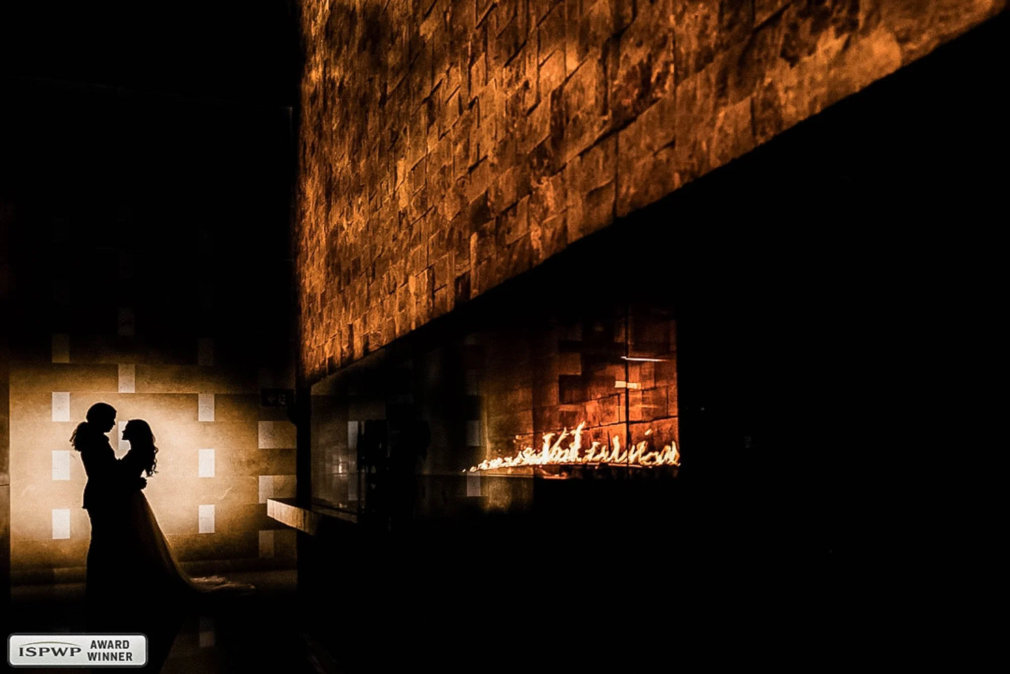 A silhouetted wedding couple, with warm lighting. In the background, there is a large brick wall illuminated by a fire, creating a cozy atmosphere.