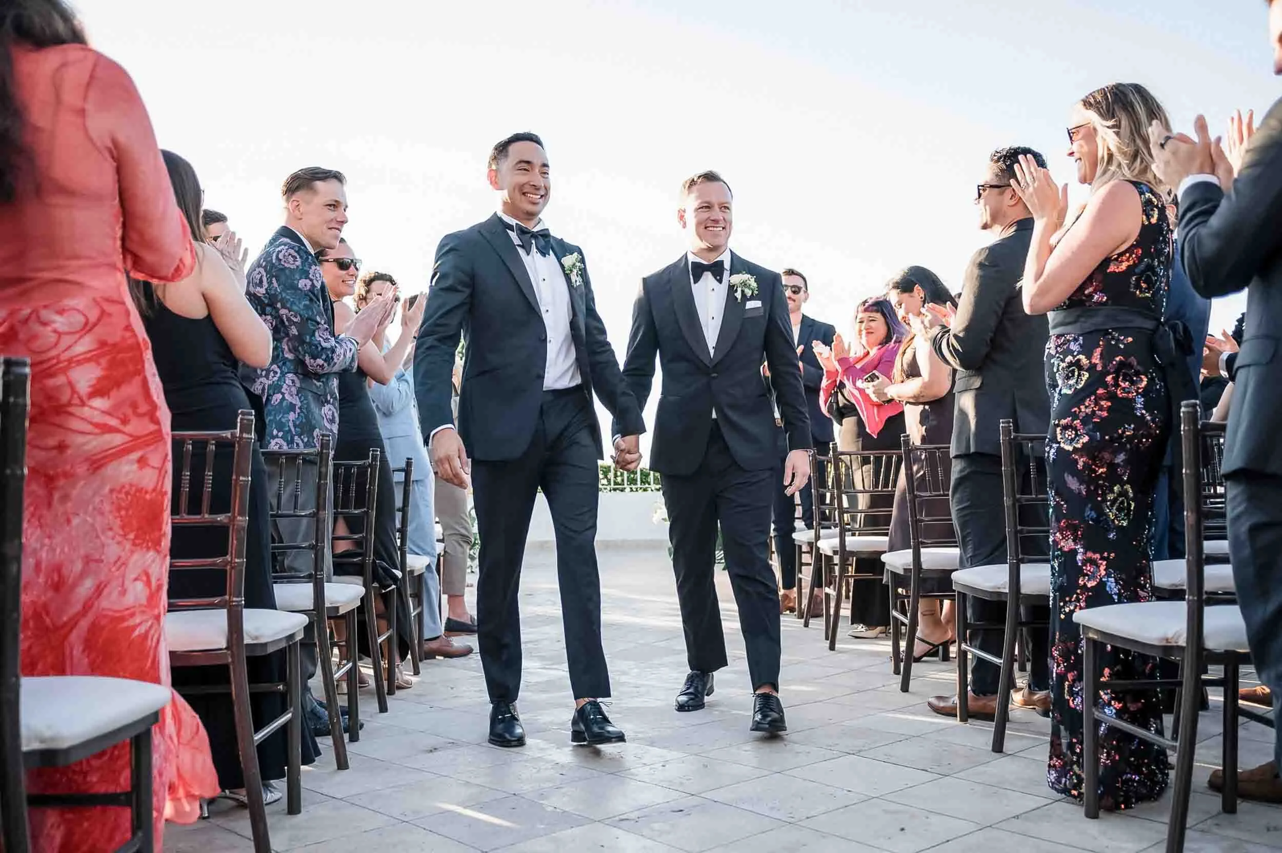 A gay wedding ceremony outdoors with two men dressed in black tuxedos holding hands, walking down the aisle surrounded by seated guests applauding and smiling.