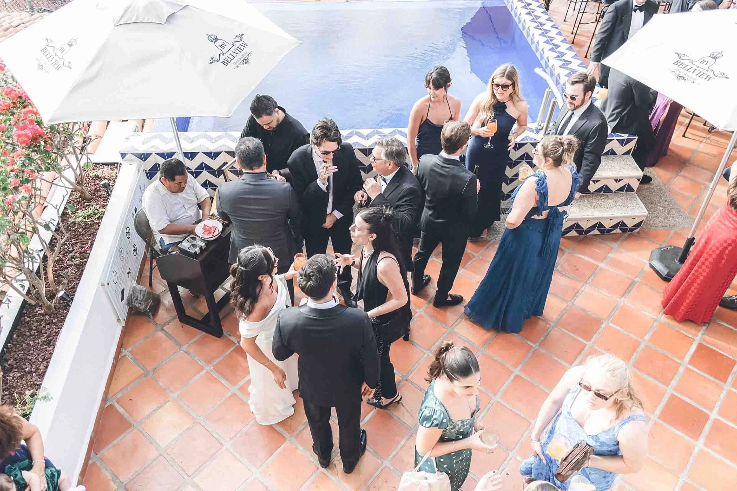 People socializing around a swimming pool at a destination wedding, some holding drinks, under umbrellas with a tiled pool edge and terracotta floor.