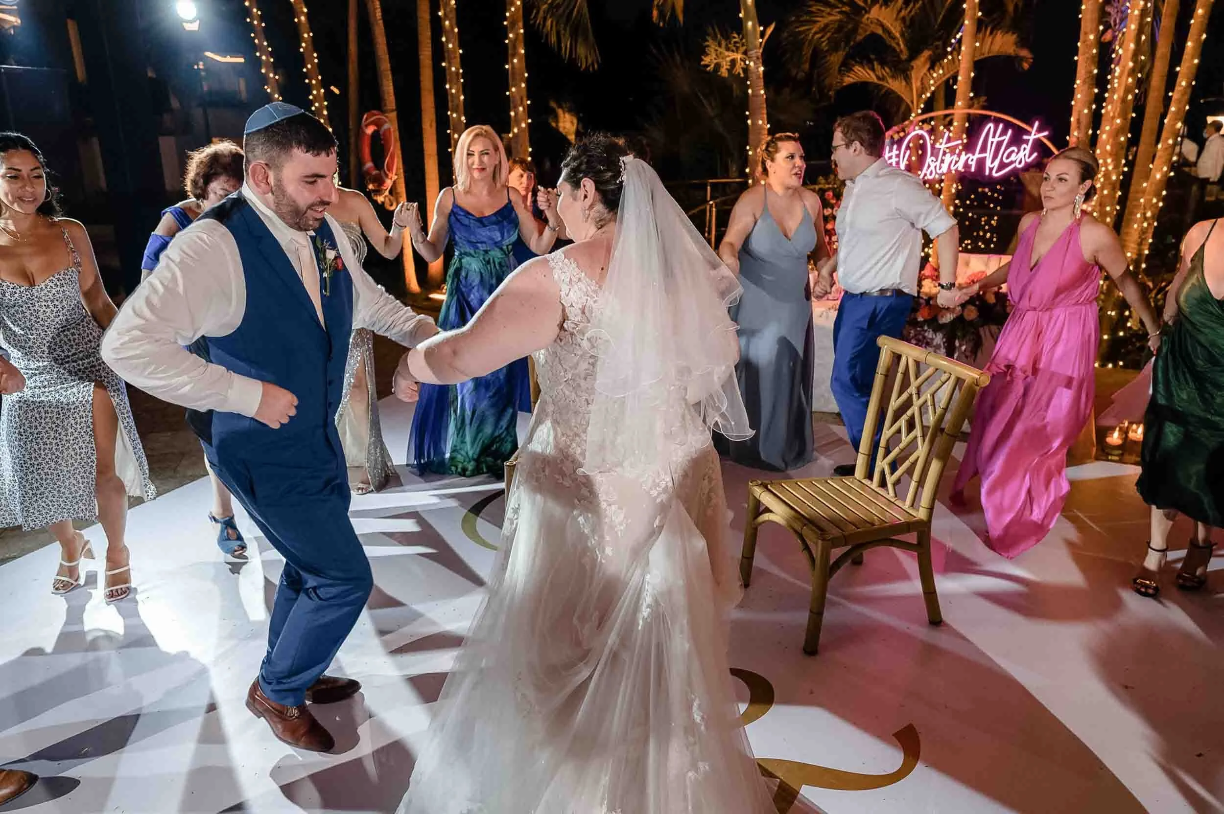 People dancing at a wedding reception under string lights and palm trees.