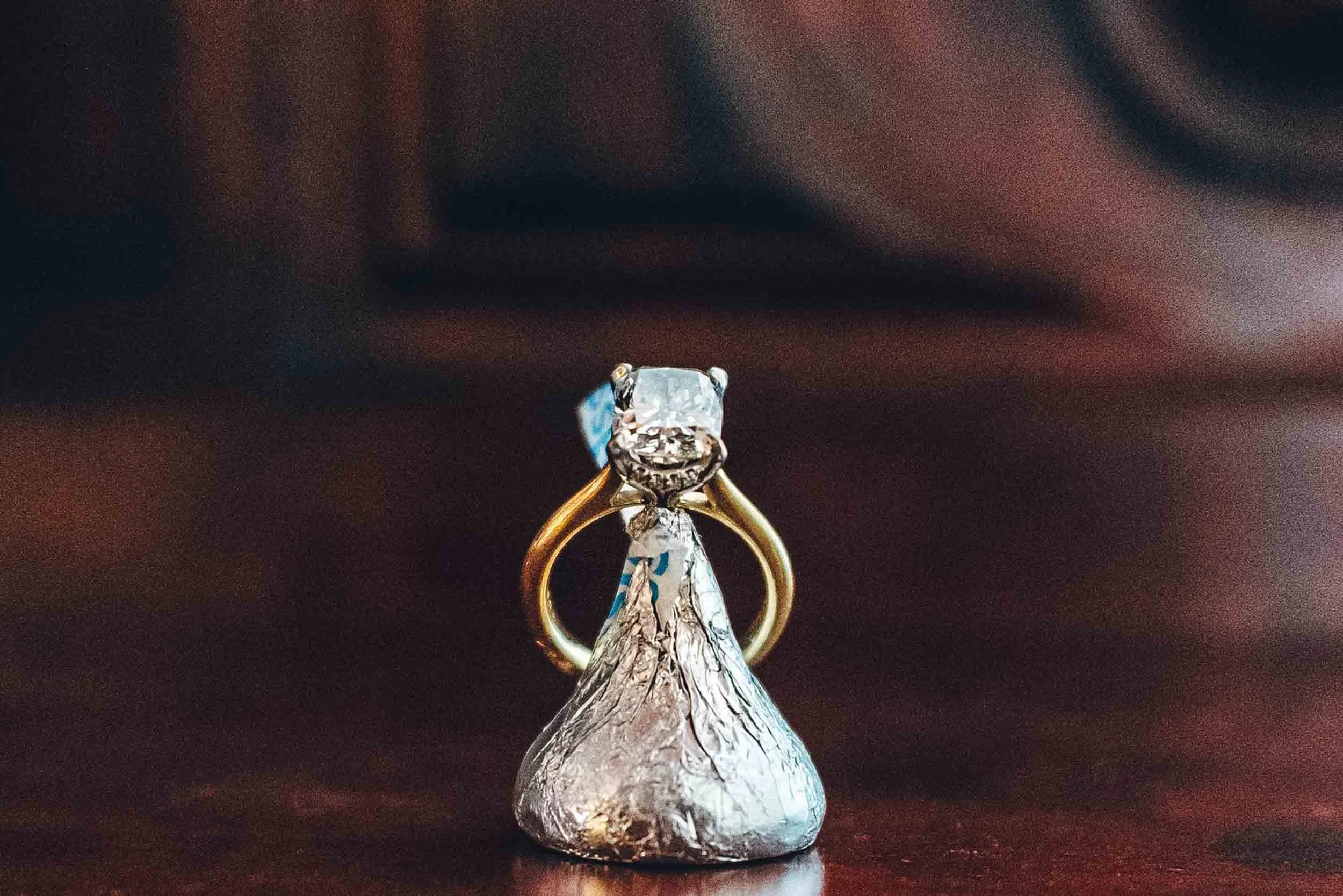 A close-up of an engagement  ring with a large diamond,  ring is placed on top of a wrapped chocolate Kiss. 