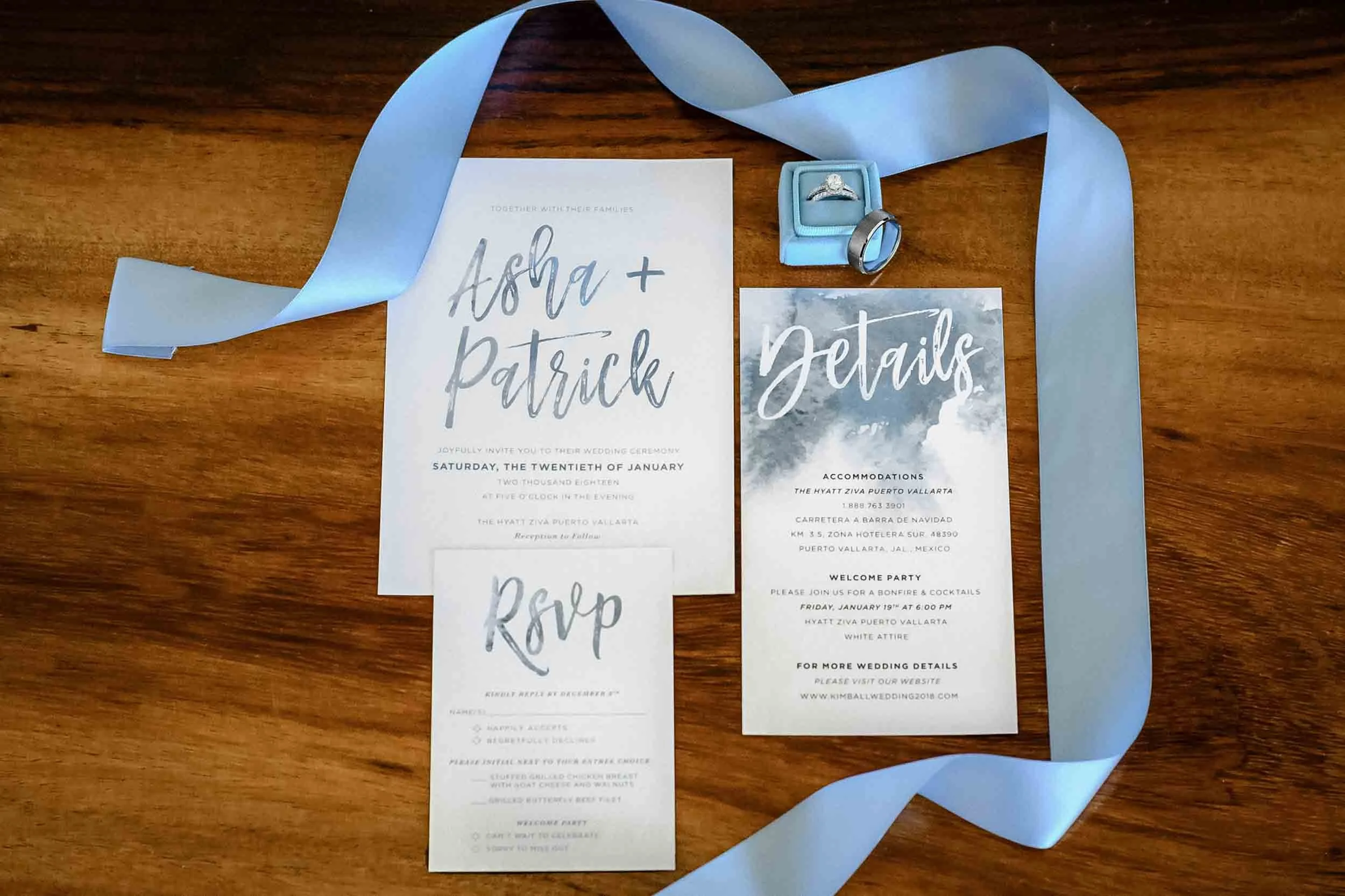 Wedding invitation, RSVP card, a silver engagement ring in a blue box, a wedding band in a silver ring holder, a blue ribbon, and a wedding details card on a wooden table.