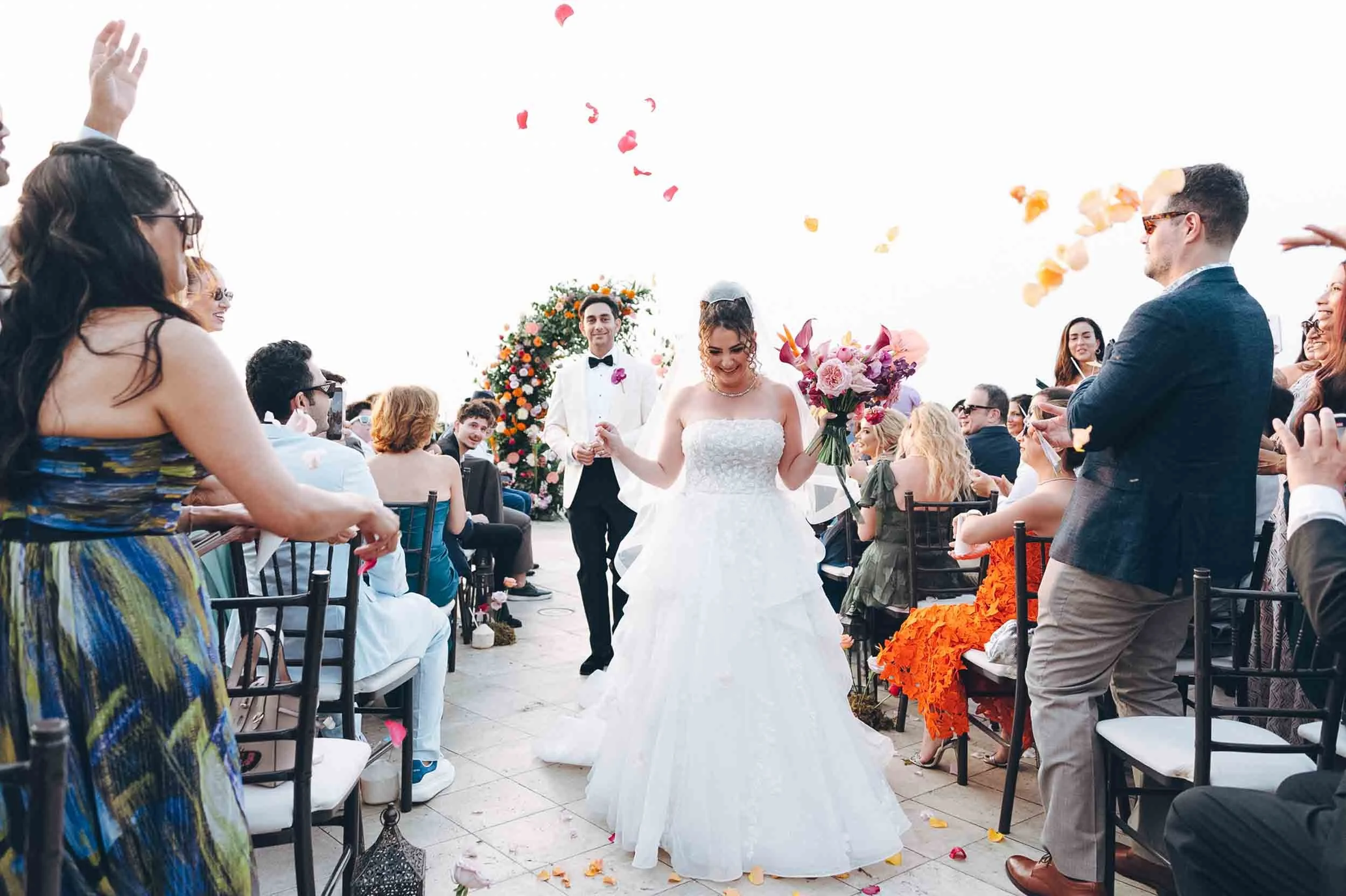 A bride and groom walking down the aisle during the exit at an outdoor wedding ceremony, with guests seated on either side and colorful flower petals being thrown in the air.