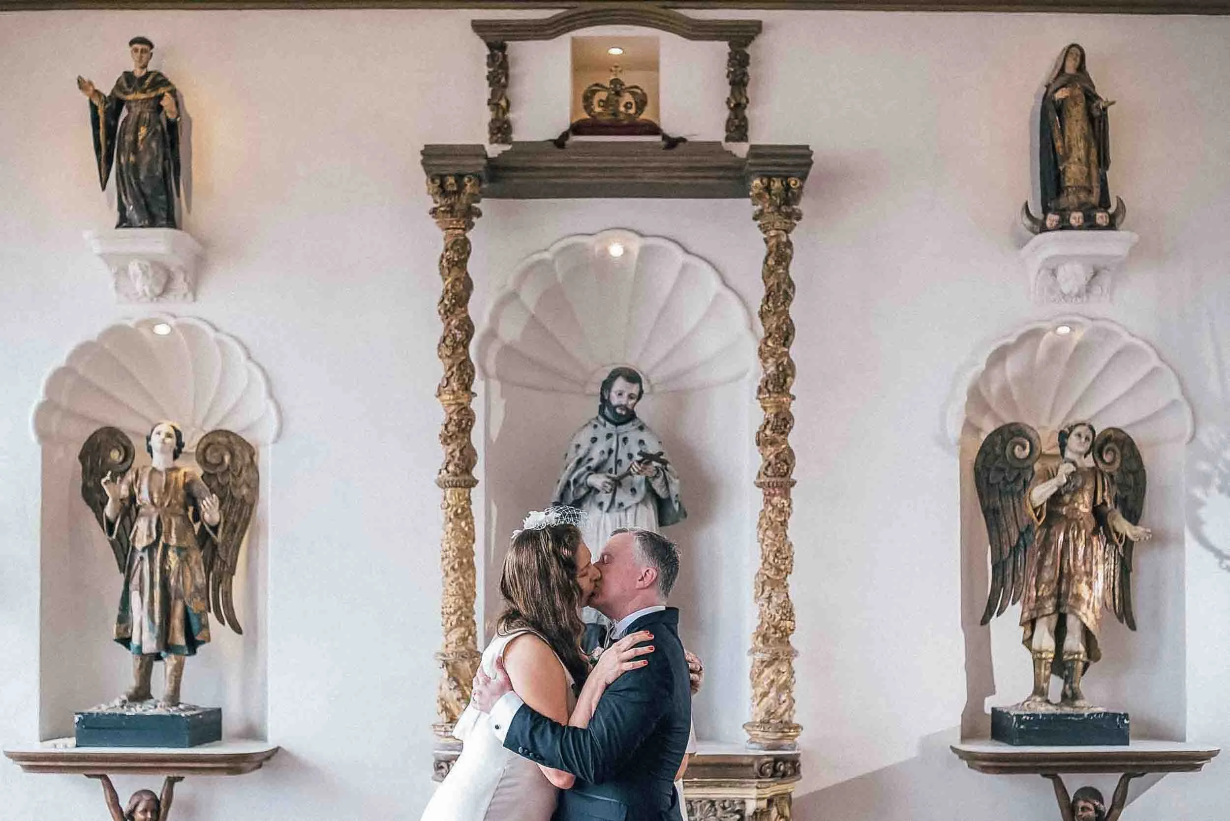 A couple kissing at their wedding ceremony in front of religious statues and artwork in venue.