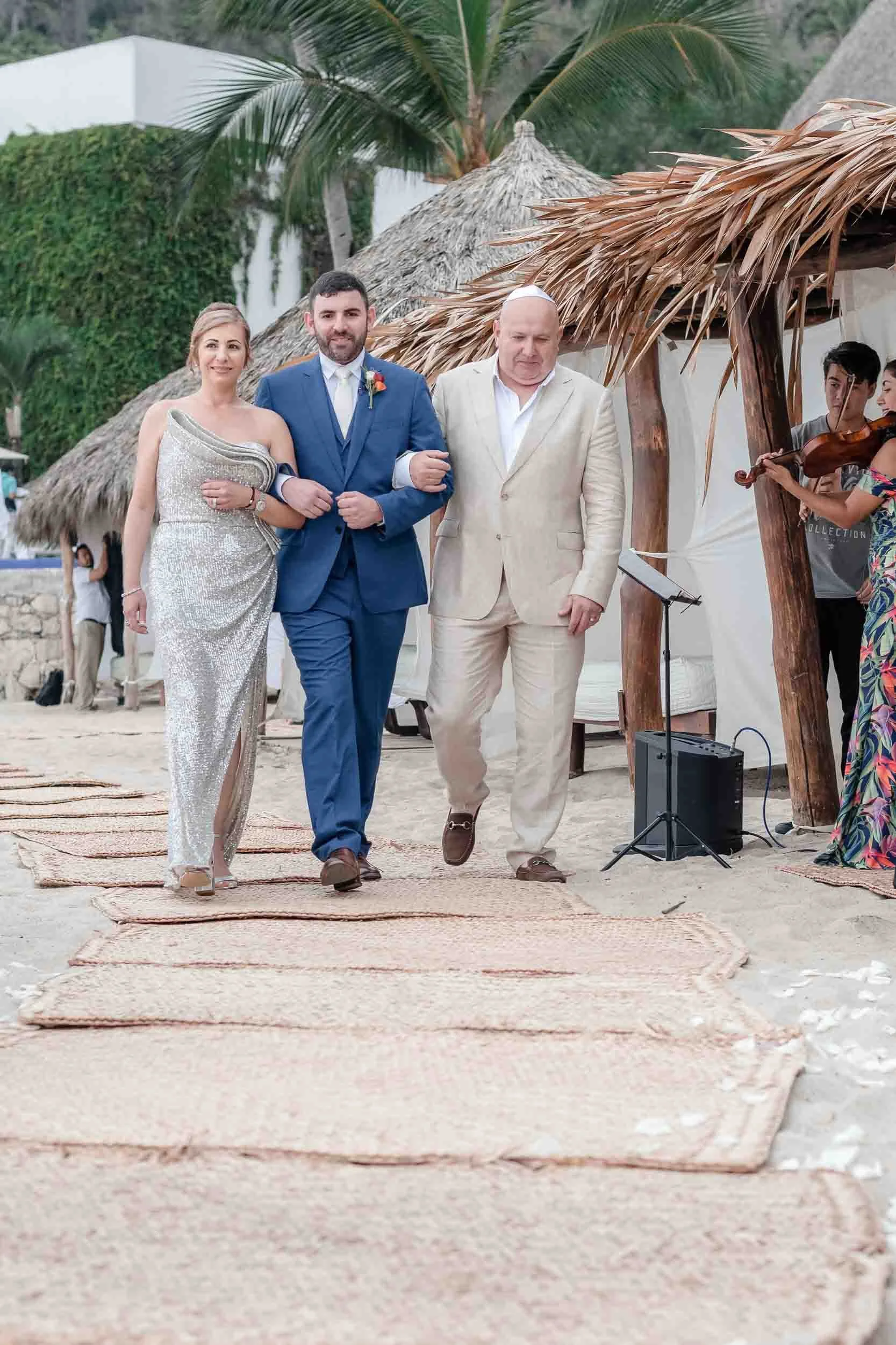 Parents walking down the aisle at a beach wedding accompanying the groom. Thatched umbrellas and palm trees are in the background.