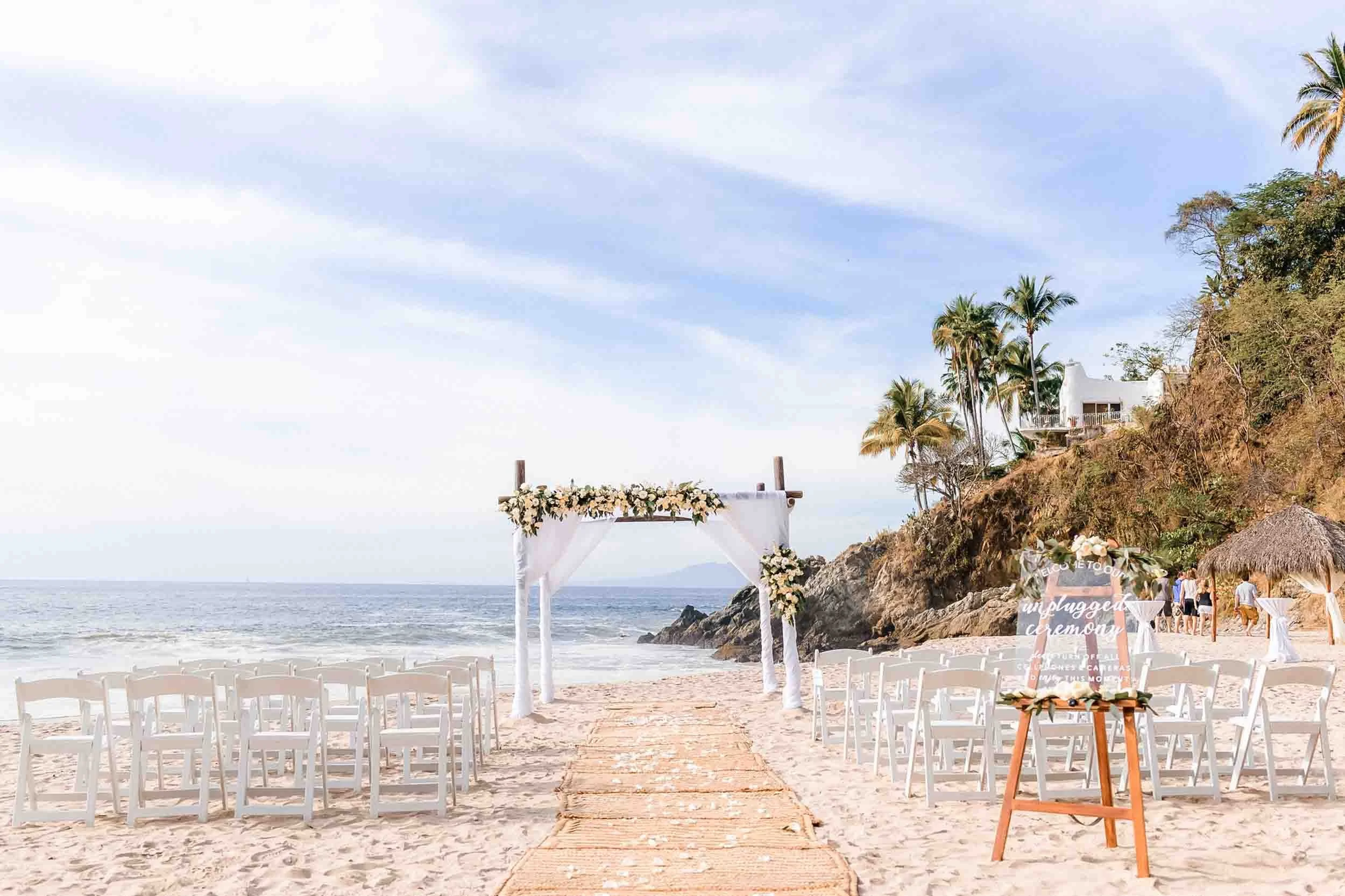 A wedding setup on a beach with white chairs arranged on both sides of a sandy aisle leading to an arch decorated with white flowers, overlooking the ocean, at the Hyat Ziva resort.