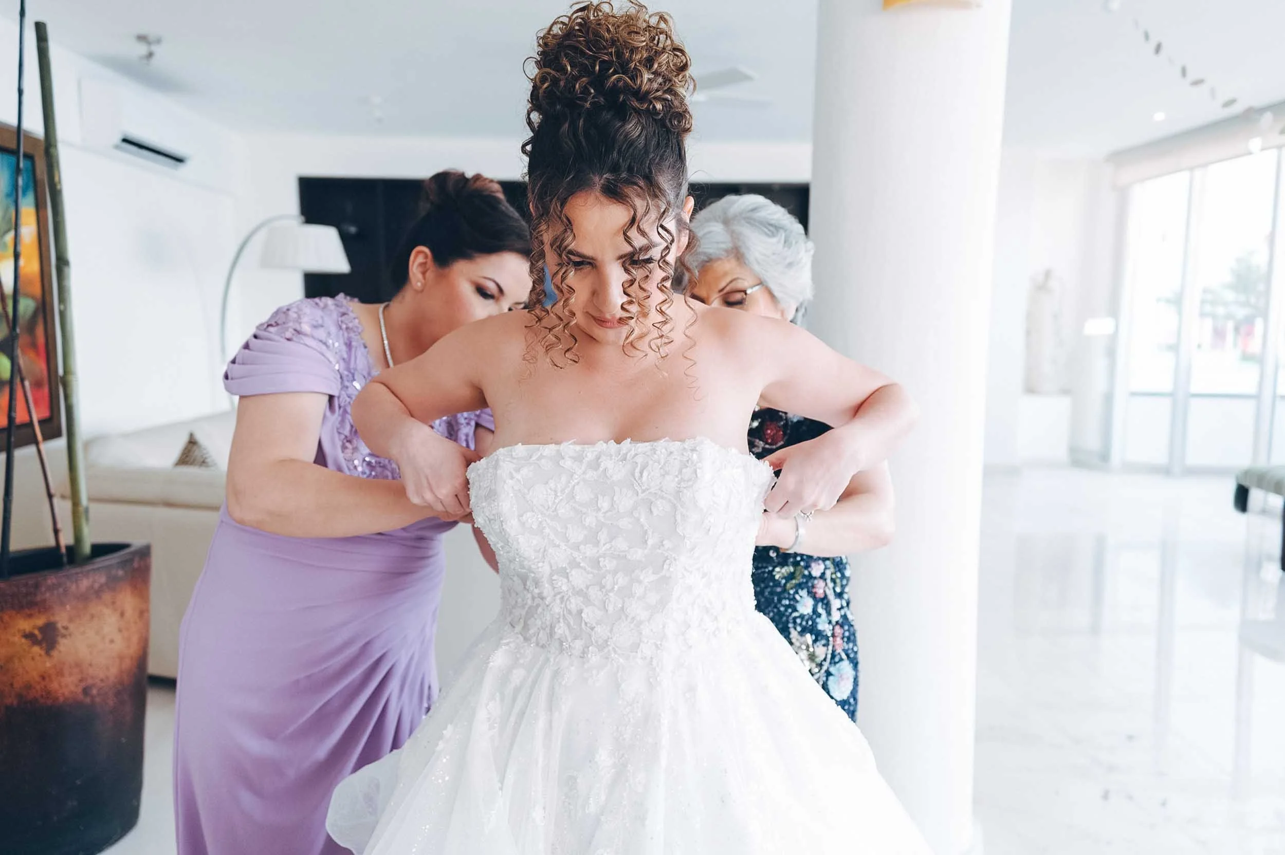 A bride in a strapless lace wedding gown is assisted by two women as she prepares for her wedding. The women are helping her adjust her dress, with one wearing a lavender dress and the other an floral dress.