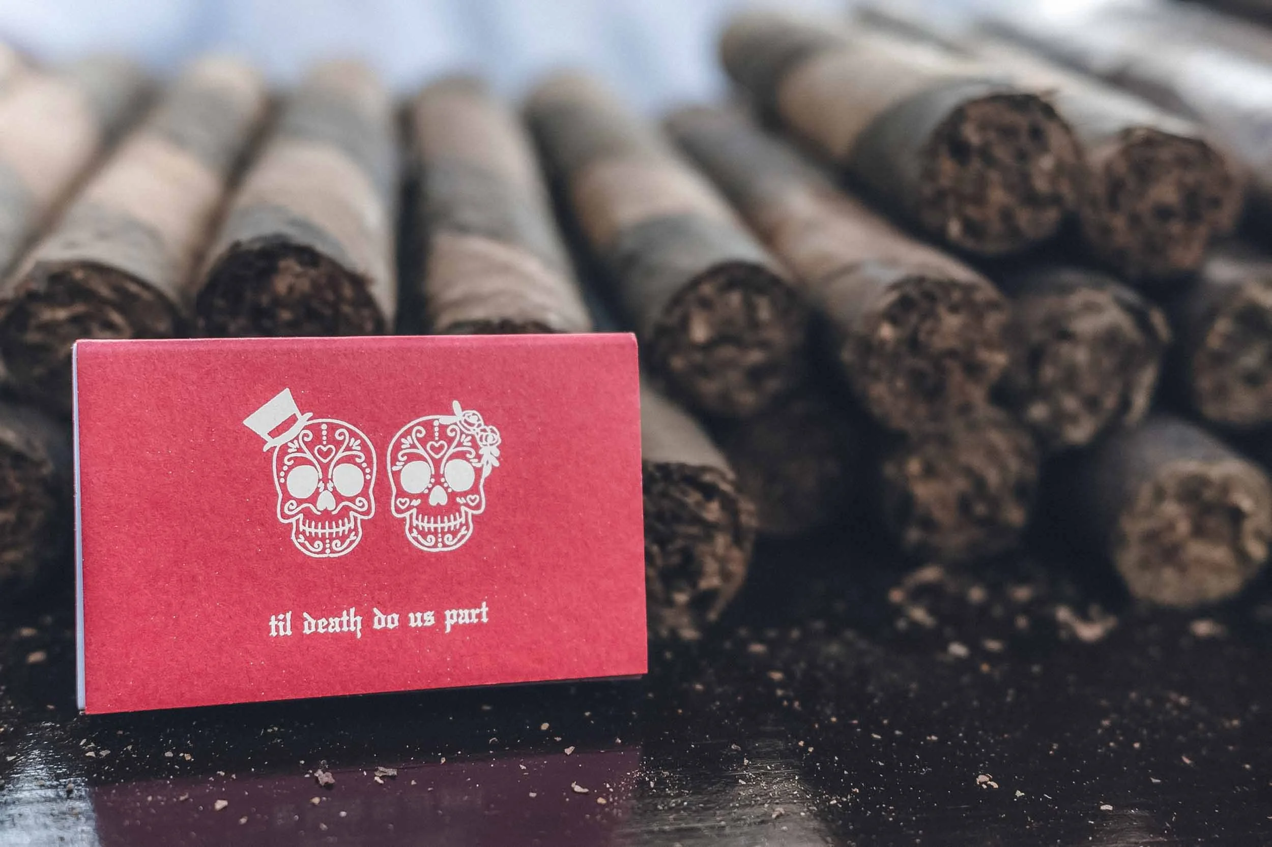 Close-up of a red matchbook keepsake with two decorated skulls on it, placed in front of a row of hand rolled cigars.